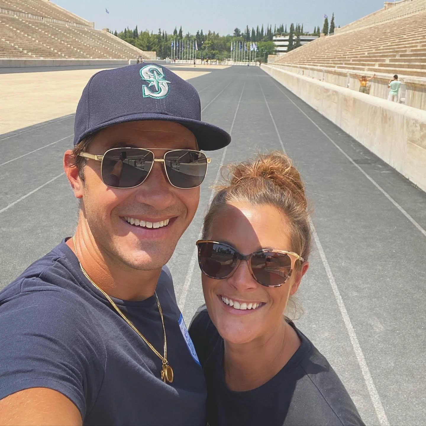 Two smiling people, a man and a woman, taking a selfie together at a football stadium with empty bleachers and flagpoles in the background on a sunny day.