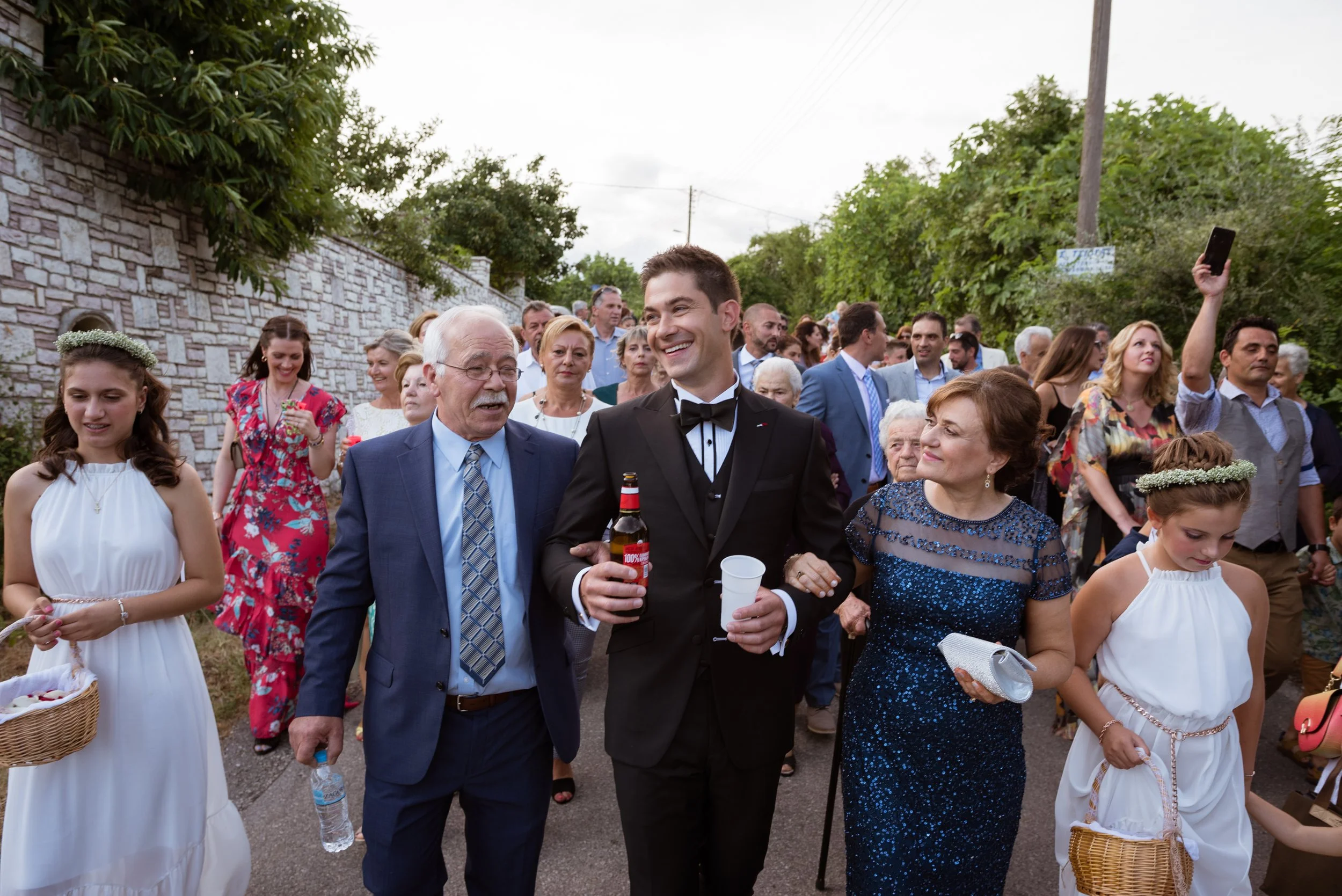People attending a celebration or wedding outdoors, with elderly and young adults dressed in formal attire, some holding drinks and a musical instrument, walking along a street lined with trees and a brick wall.