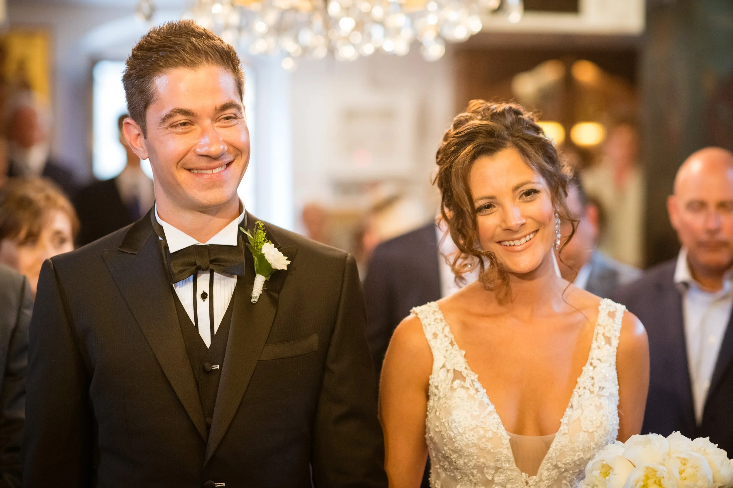 A smiling man in a black tuxedo and a woman in a white lace wedding dress standing together at a wedding ceremony, with guests and a chandelier in the background.
