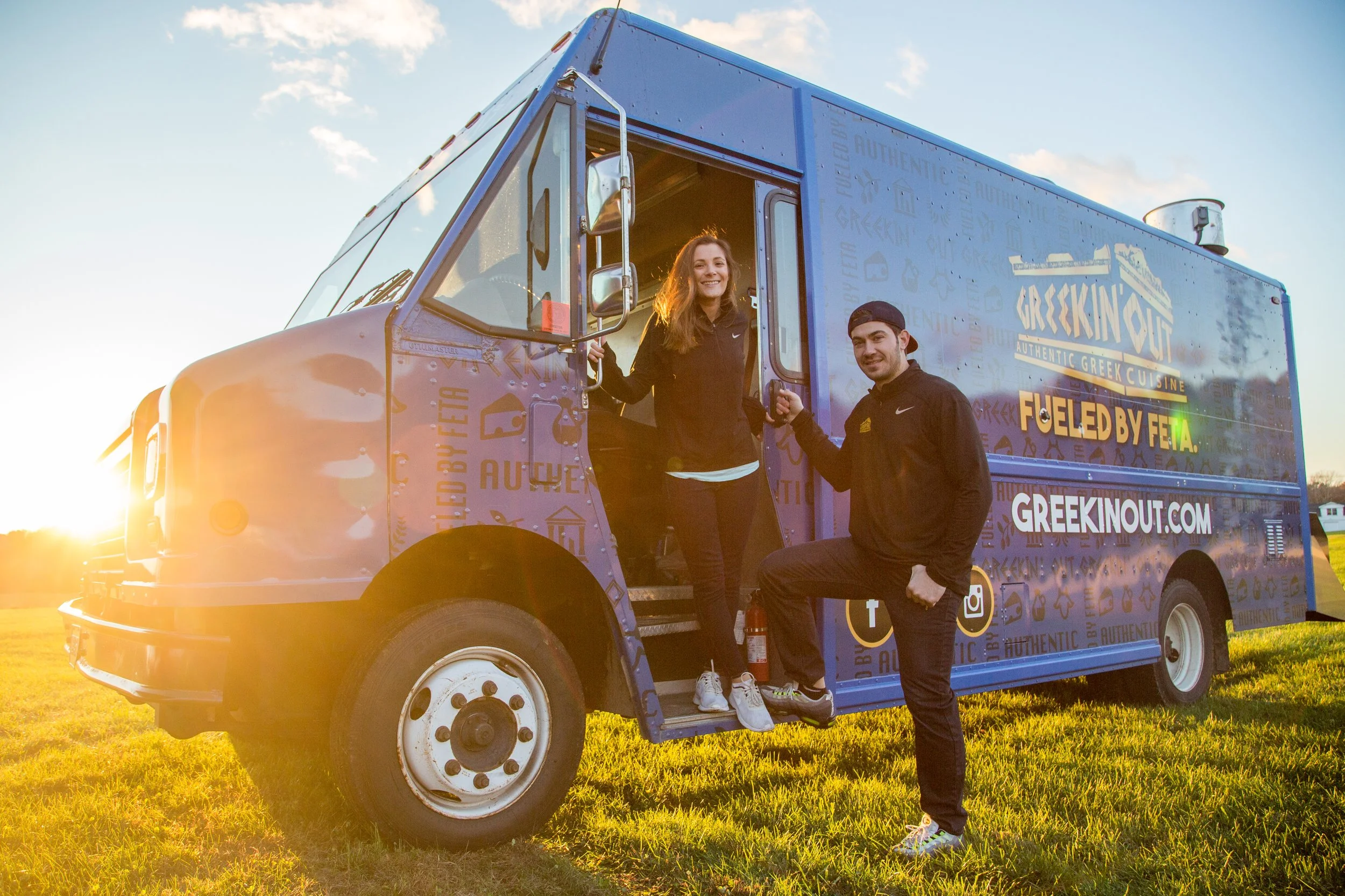 A woman and a man standing outside a blue food truck at sunset. The woman is coming out of the truck, and the man is kneeling beside the door. The food truck has text and logos on it related to Greek cuisine and social media icons.