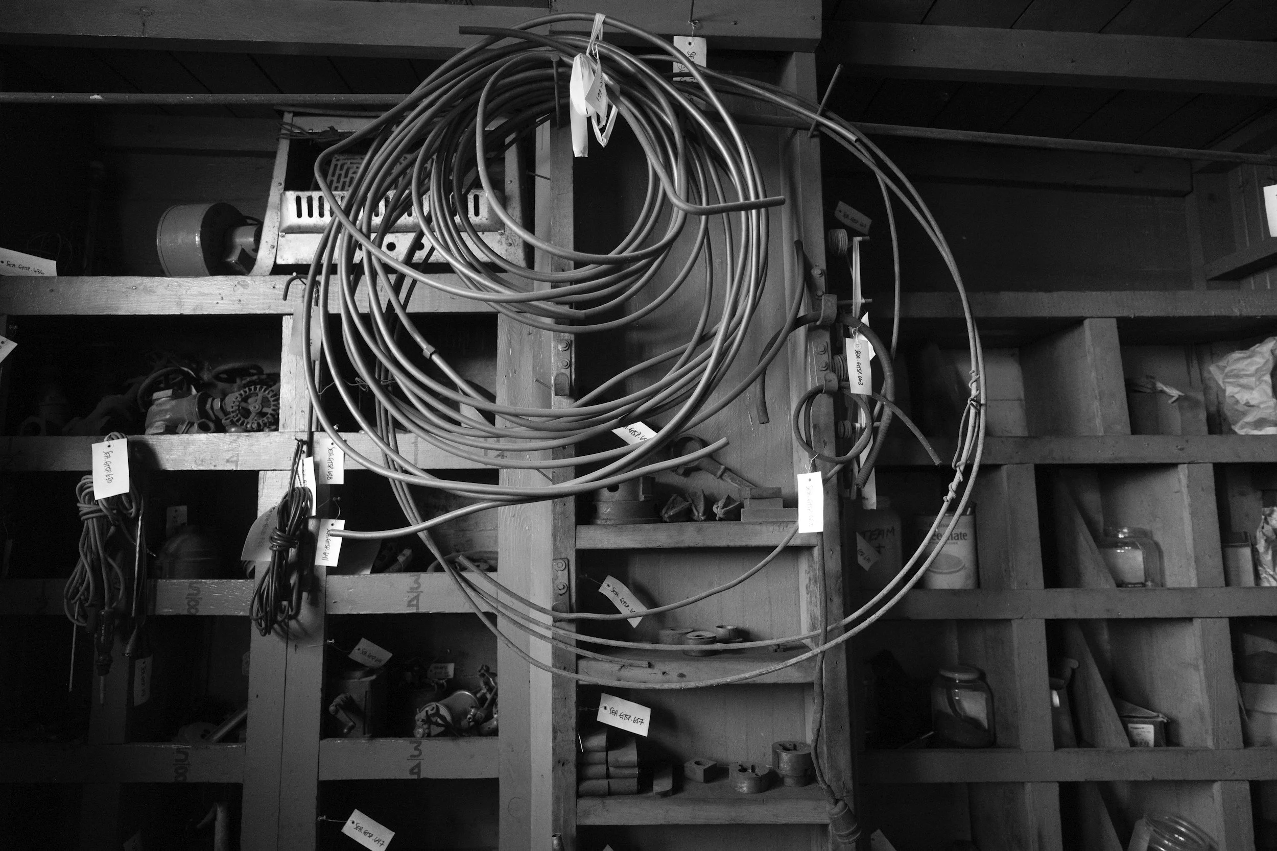 A cluttered wooden storage shelf with various tools and equipment in the background, including coiled cables hanging on the side in the foreground.