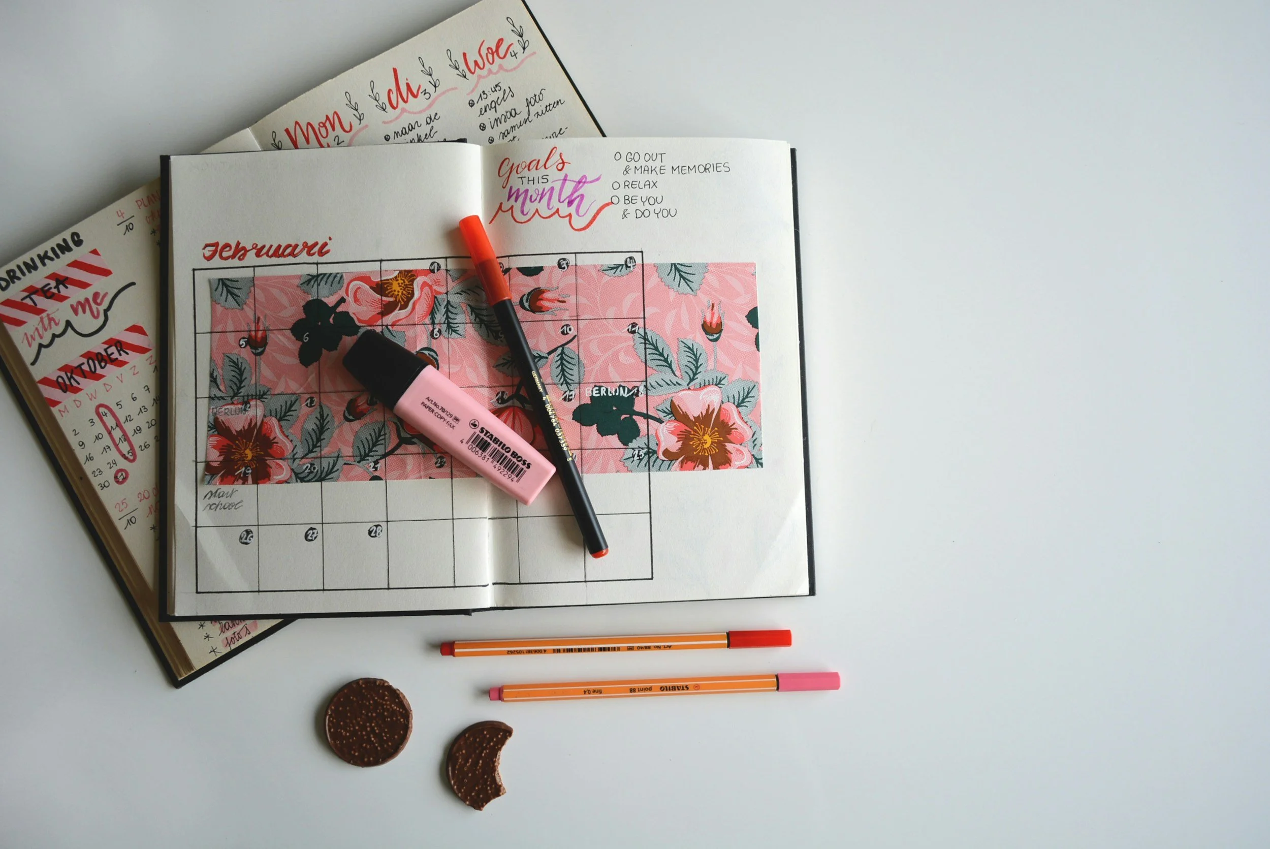 Open planner with February calendar, floral decorations, markers, a sticky note, two chocolate cookies, and a blank notebook on a white surface