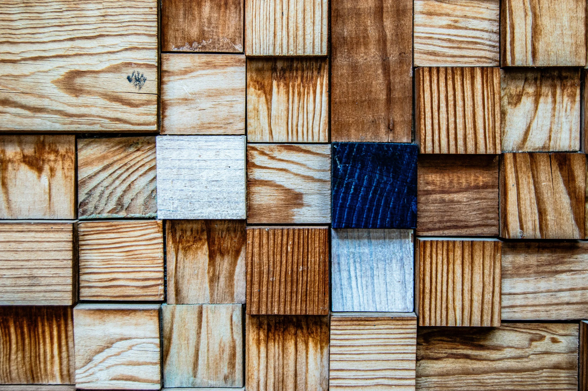 Close-up view of a wall made of wooden blocks arranged in a grid pattern, with some blocks darker or painted, creating a mosaic-like appearance.