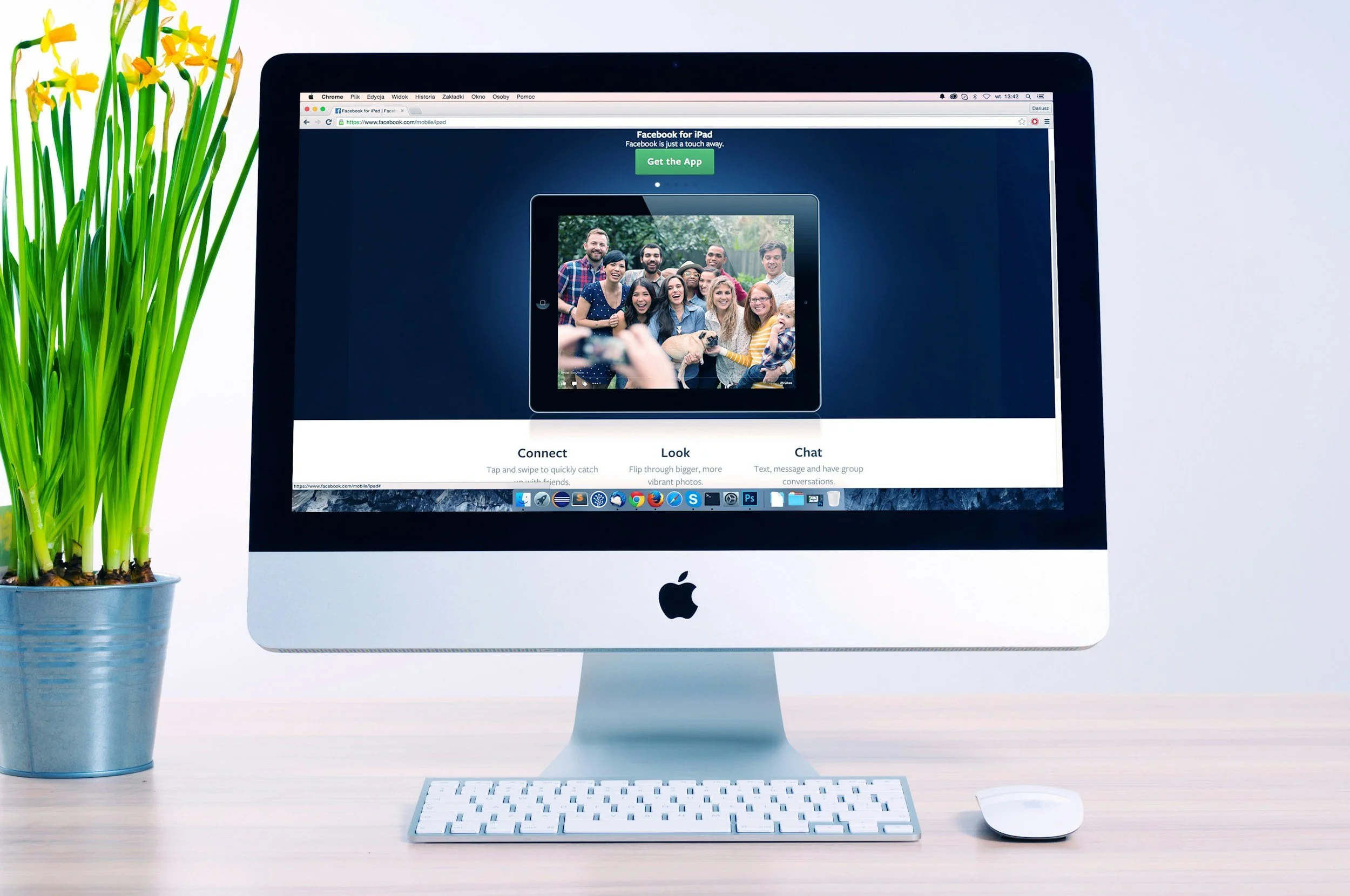 An Apple iMac on a desk with a keyboard and mouse, displaying a Facebook page about a photo of a group of people, beside a potted plant with green leaves and yellow flowers.