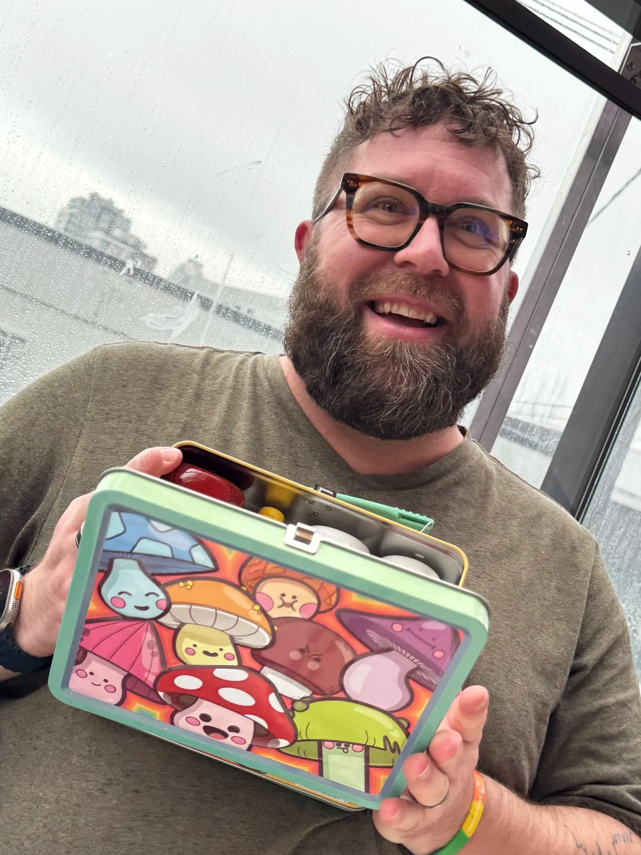 A smiling man with glasses and a beard holding a colorful tin box with cartoon mushroom characters inside, standing in front of a rainy window.