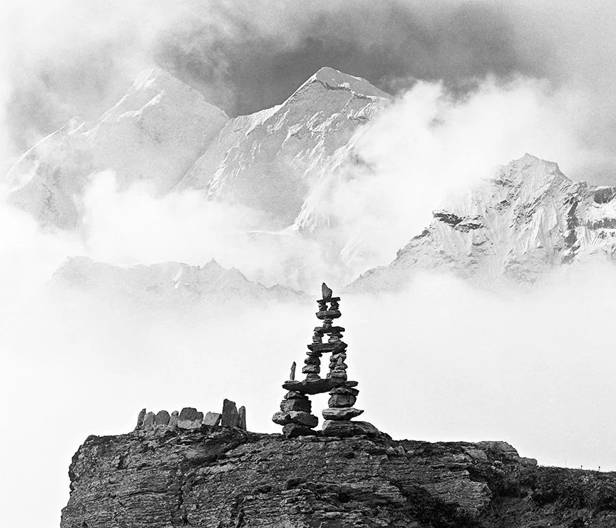 Stacked rocks forming a cairn on a rocky surface with snow-capped mountain peaks and clouds in the background.