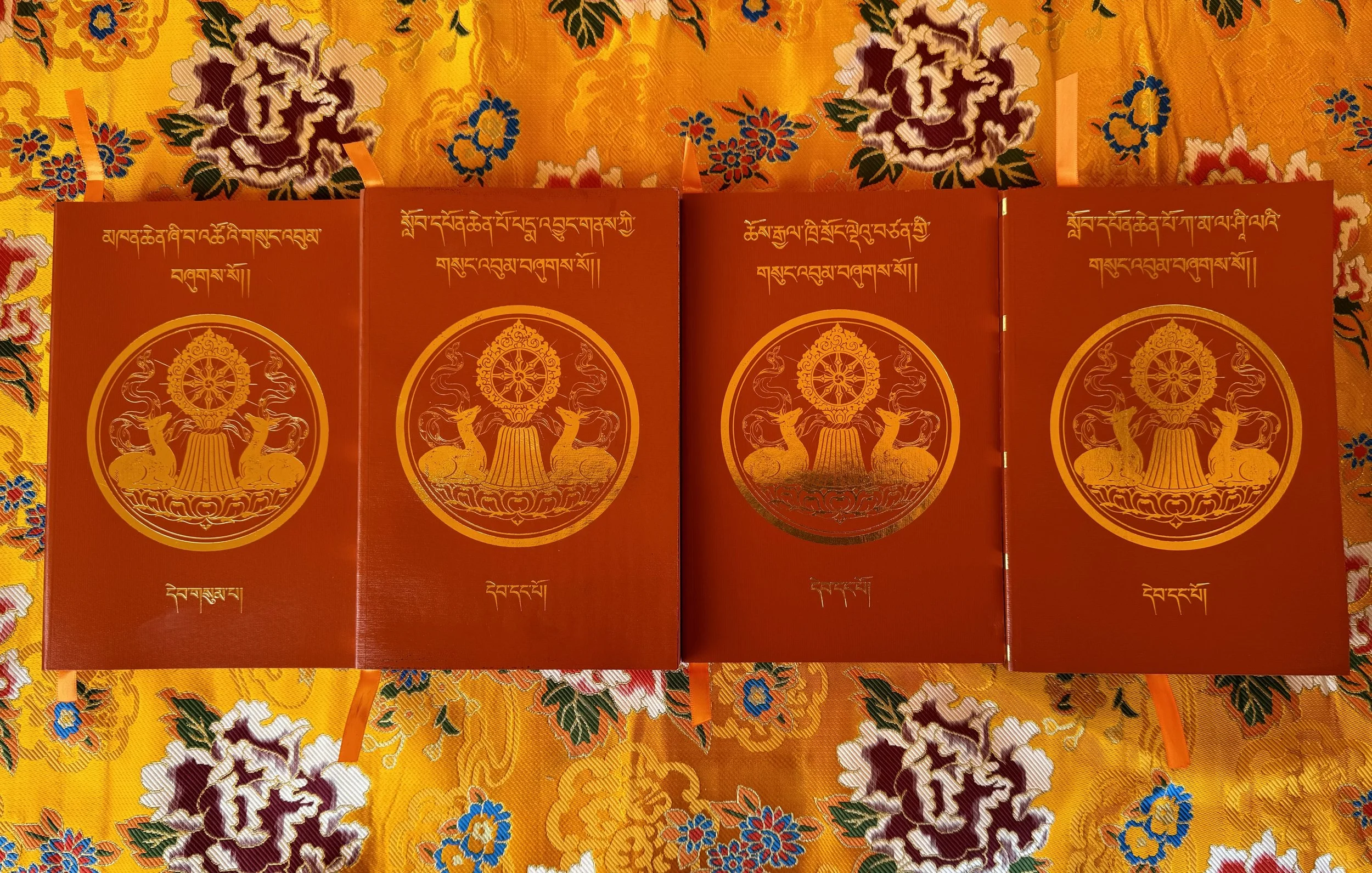 Four red books with gold Tibetan script and emblem of two deer on a yellow floral cloth background.