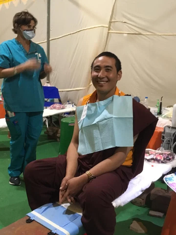 A man wearing traditional Buddhist monk robes and a watch, sitting on a chair with a paper bib over his chest, smiling after receiving dental care. A healthcare worker wearing scrubs and a face mask standing nearby in a medical setting.