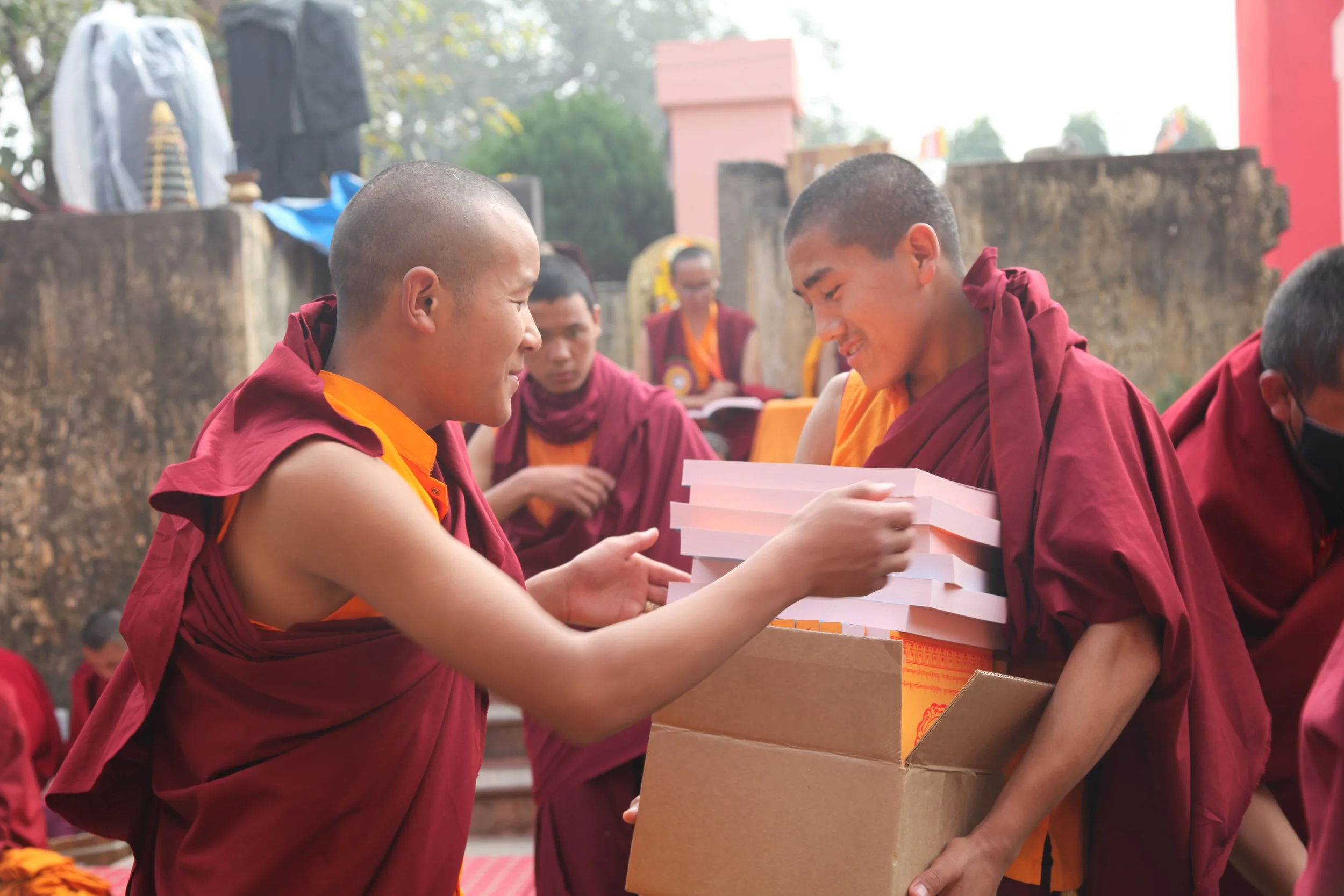 A Buddhist monk in maroon robes receiving a stack of books from a fellow monk during a ceremony outdoors.