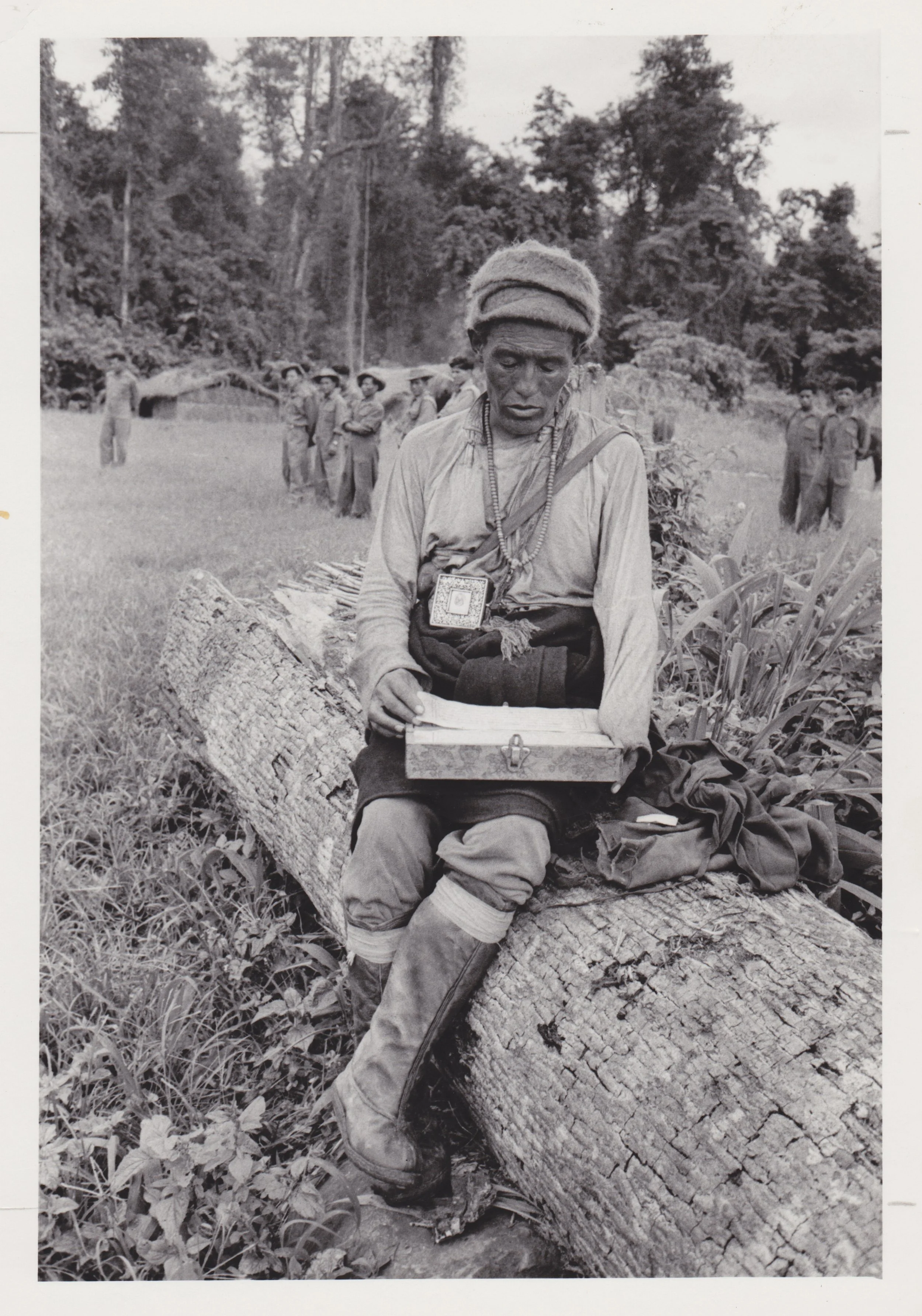 Tibetan refugee sitting on a fallen tree with a box on their lap, while a group of people stand in the background in a rural outdoor setting.