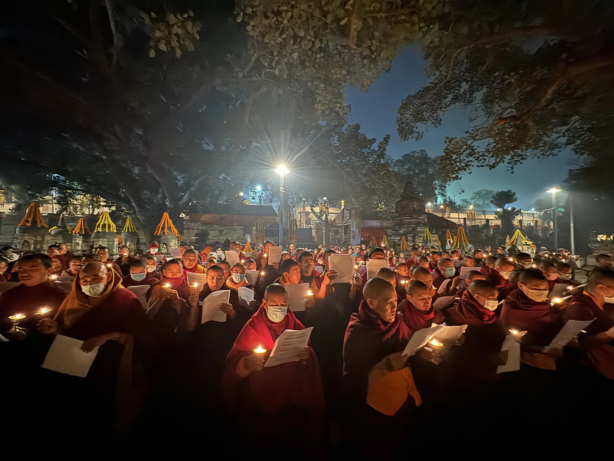 A large group of monks holding candles and papers, participating in a nighttime prayer or ceremony outdoors under large trees, with decorated structures and lights in the background.