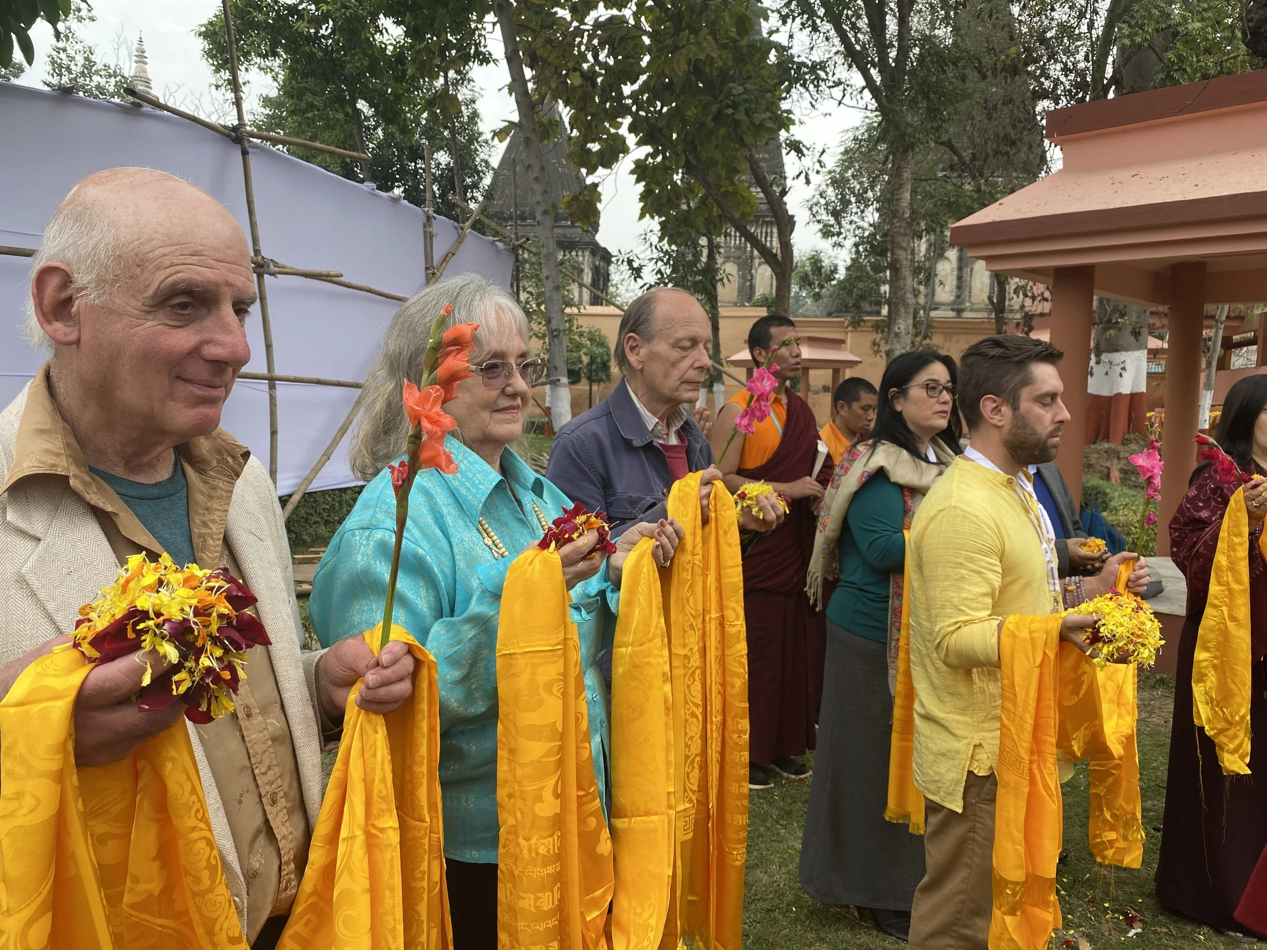 A group of people participating in a religious or cultural ceremony outdoors, holding flowers and wearing yellow scarves, with temple structures and trees in the background.