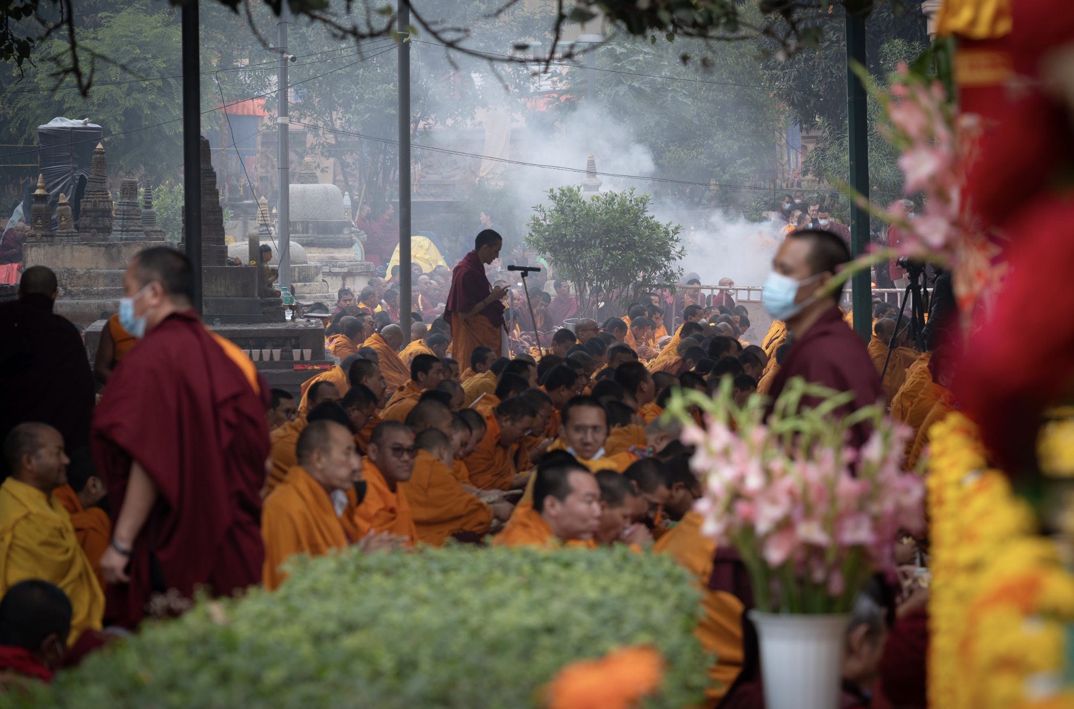 A large gathering of Buddhist monks in orange and maroon robes, many wearing face masks, seated outdoors during a religious ceremony. Smoke and incense are visible in the background, along with a monk standing and speaking into a microphone.