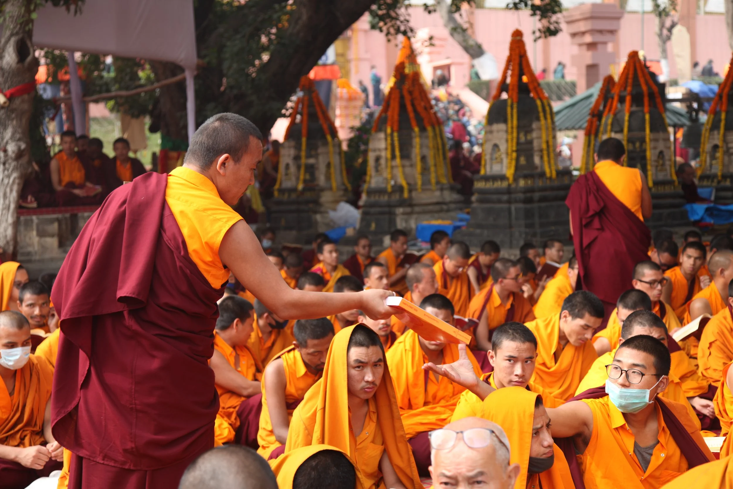 Monks in orange and maroon robes participating in a religious ceremony outdoors, some wearing face masks. A monk is handing out a book to another monk, with several structures decorated with flowers in the background.