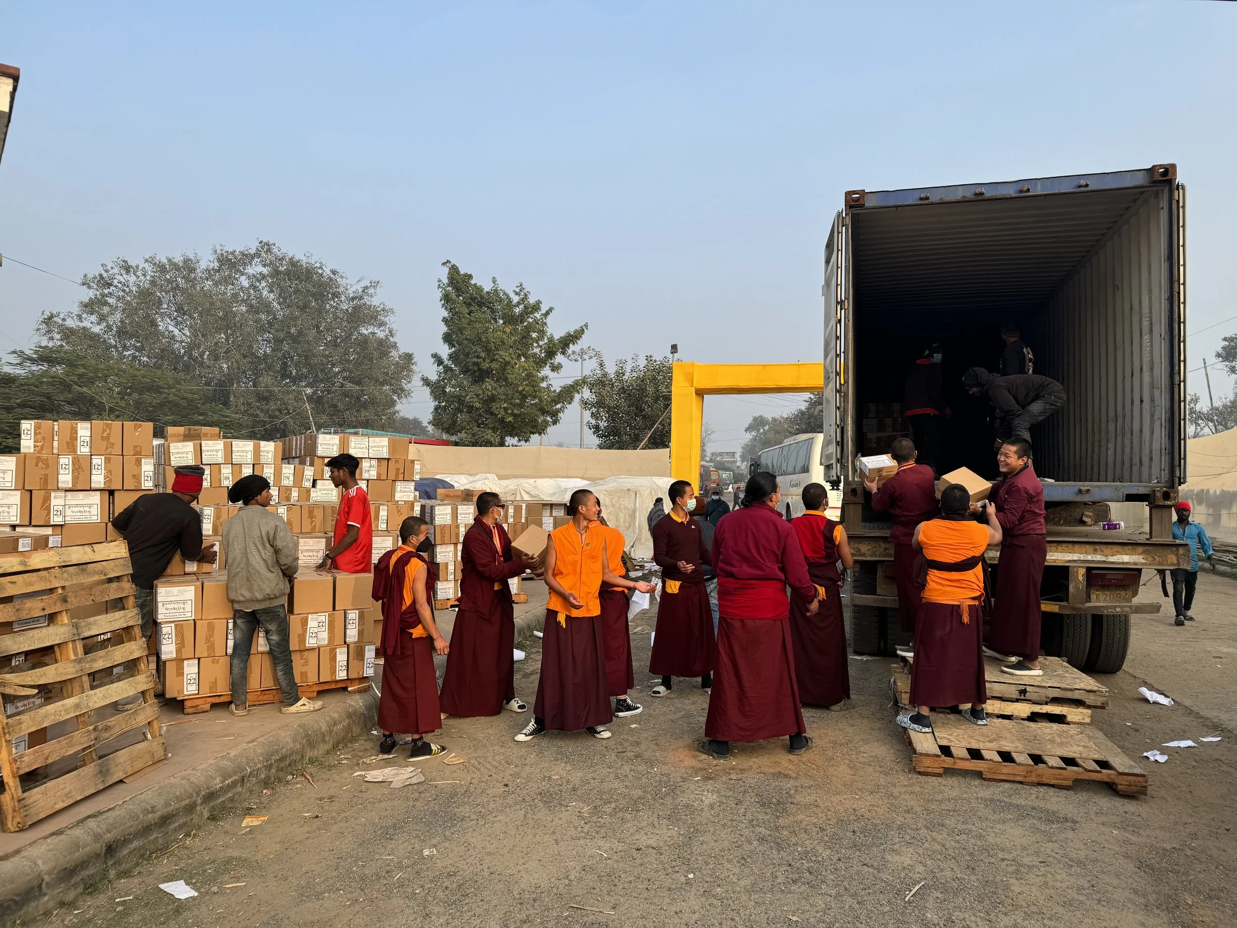 People loading boxes from a truck with a large number of boxes stacked on pallets nearby. Many individuals are participating in the unloading process, some with masks, in an outdoor setting with trees in the background.