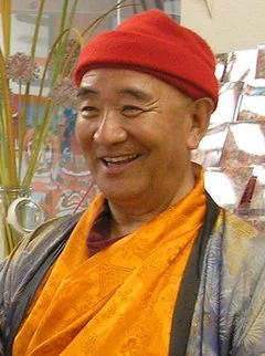 A smiling man wearing a red beanie, orange robe, and a patterned shirt, standing in front of a colorful artistic background with plants.