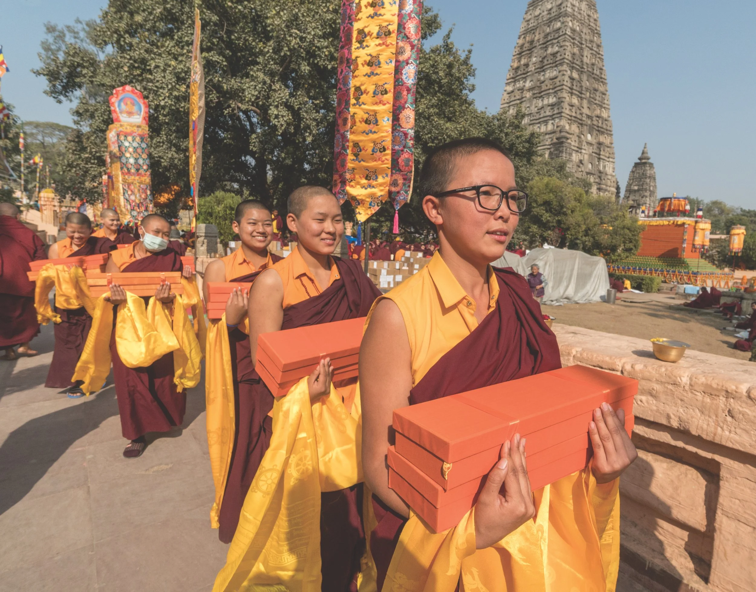 Group of young Buddhist nuns dressed in maroon and yellow robes, carrying books, walking in a line at a temple complex with ancient pagodas, colorful prayer flags, and flowering trees in the background. 