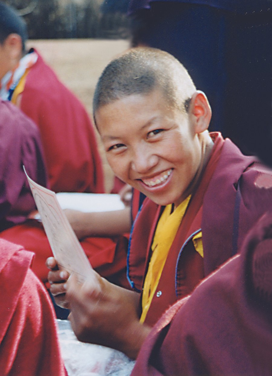 A smiling nun wearing a yellow shirt and a maroon jacket, holding a piece page of a loose-leafed book.