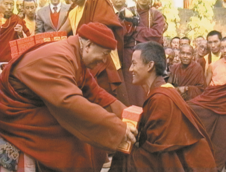 Tarthang Rinpoche handing a book to a young monk, surrounded by a group of monks in robes, outdoors during daytime.