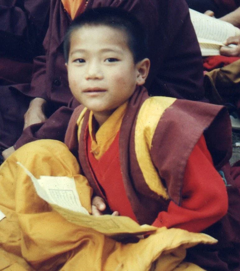 A young boy in traditional monk robes sitting with prayer beads, surrounded by other monks in similar attire.