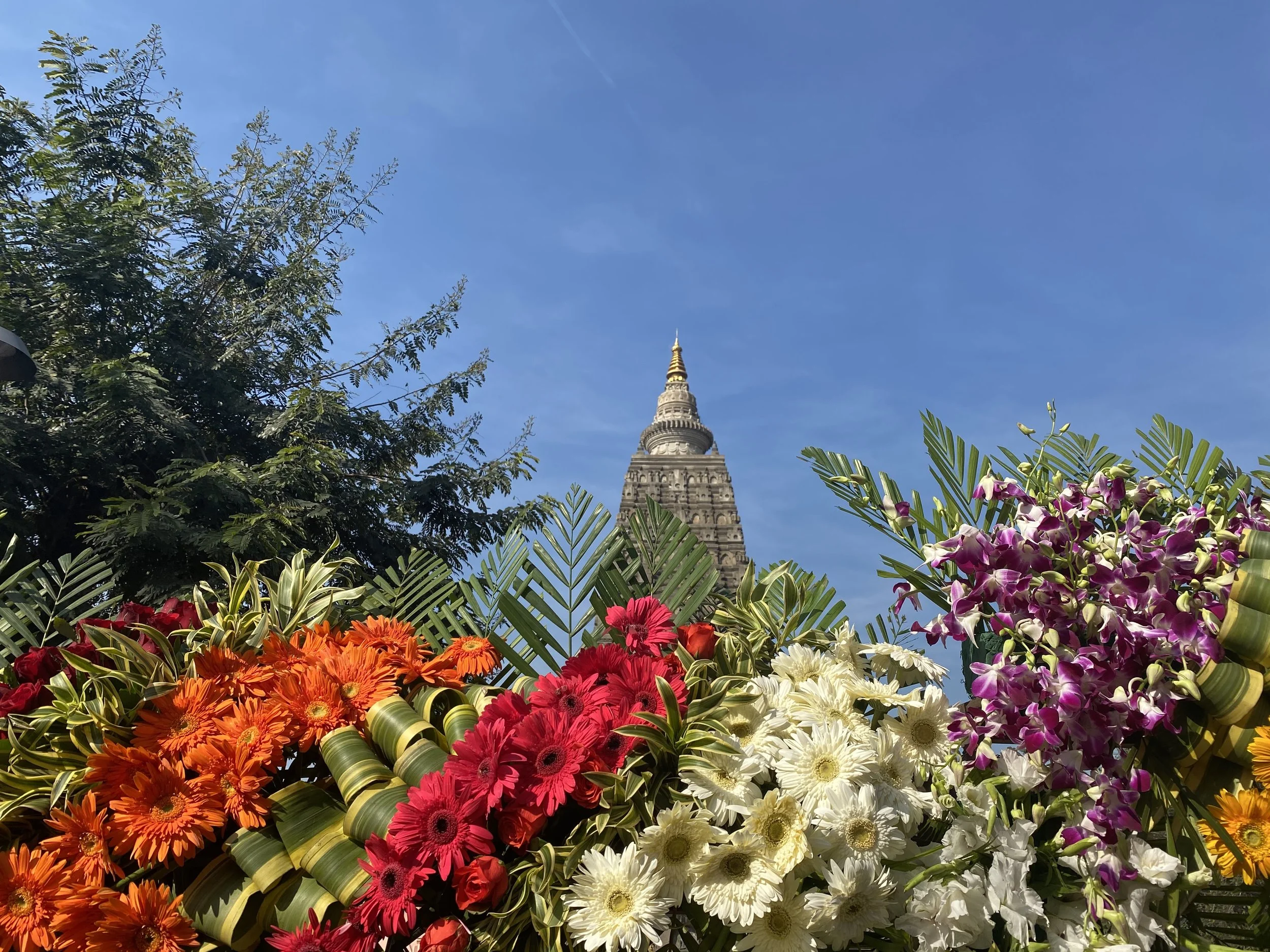 Colorful flowers including orange, red, white, and purple blooms in the foreground with the Mahabodhi temple in the background under a clear blue sky.