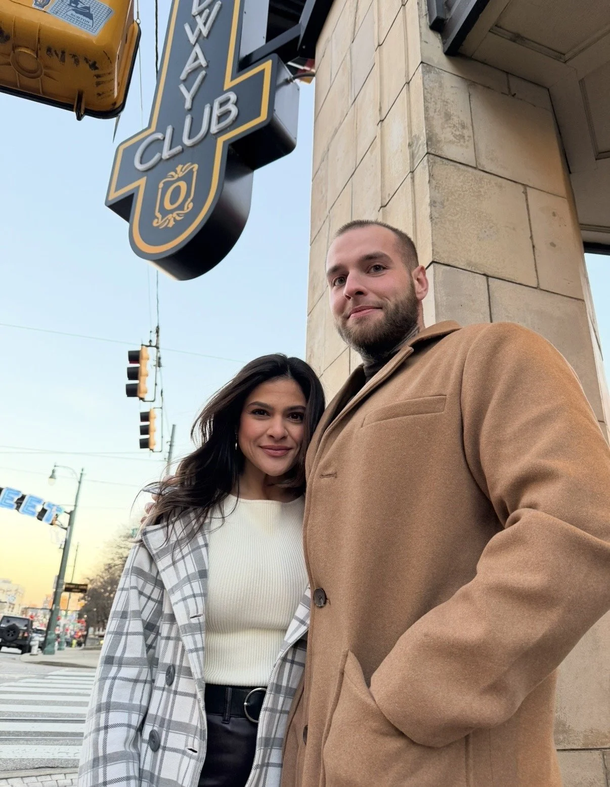 Two people, a woman and a man, standing outside near a building with a sign that reads 'WAY CLUB.' The woman has long dark hair and is wearing a white top and a plaid jacket, while the man has a beard and is wearing a tan coat. They are looking at the camera with a city street background, including traffic lights and other buildings.