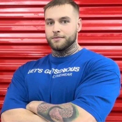 Man with short hair and beard, wearing a blue t-shirt with white text, standing in front of a red corrugated metal background.
