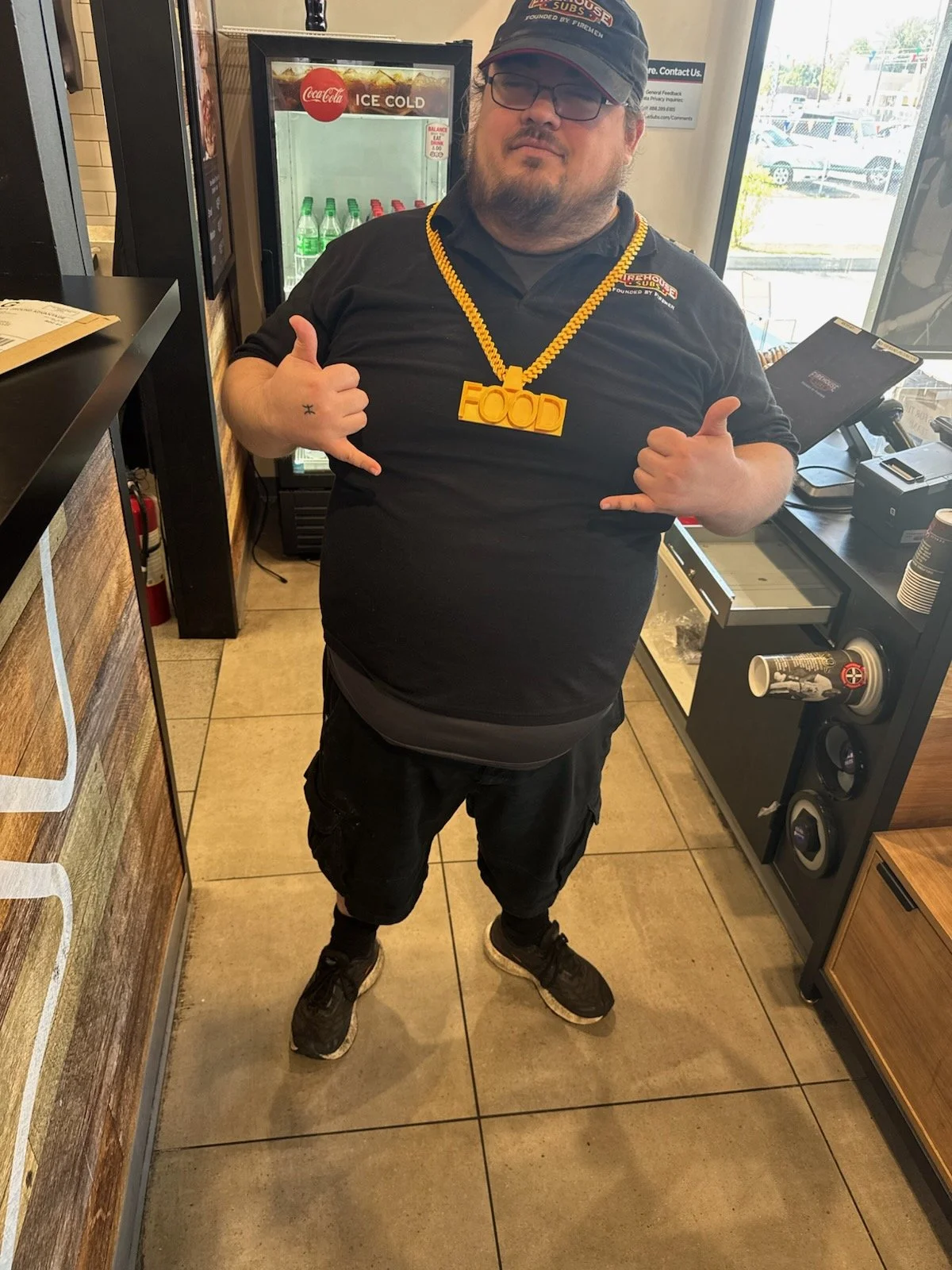 A man standing inside a fast-food restaurant, wearing a black uniform, glasses, and a red cap. He has a necklace with a large yellow "FOOD" pendant and is making a hand gesture with both hands. There is a drink machine and a counter beside him, with a window showing cars outside.