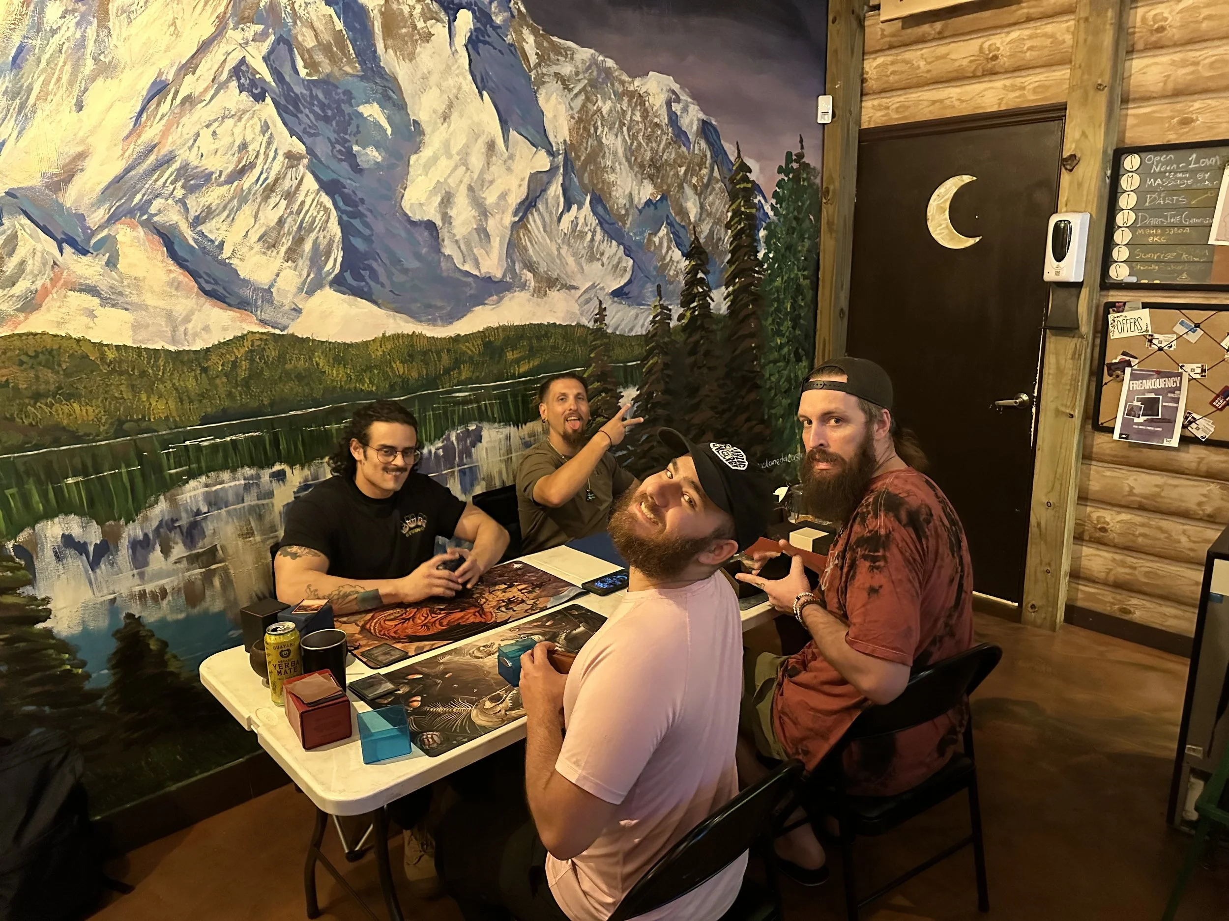 Four men sitting around a table playing a card game in a room with mountain and forest mural on the wall, and a black door with a moon cutout.