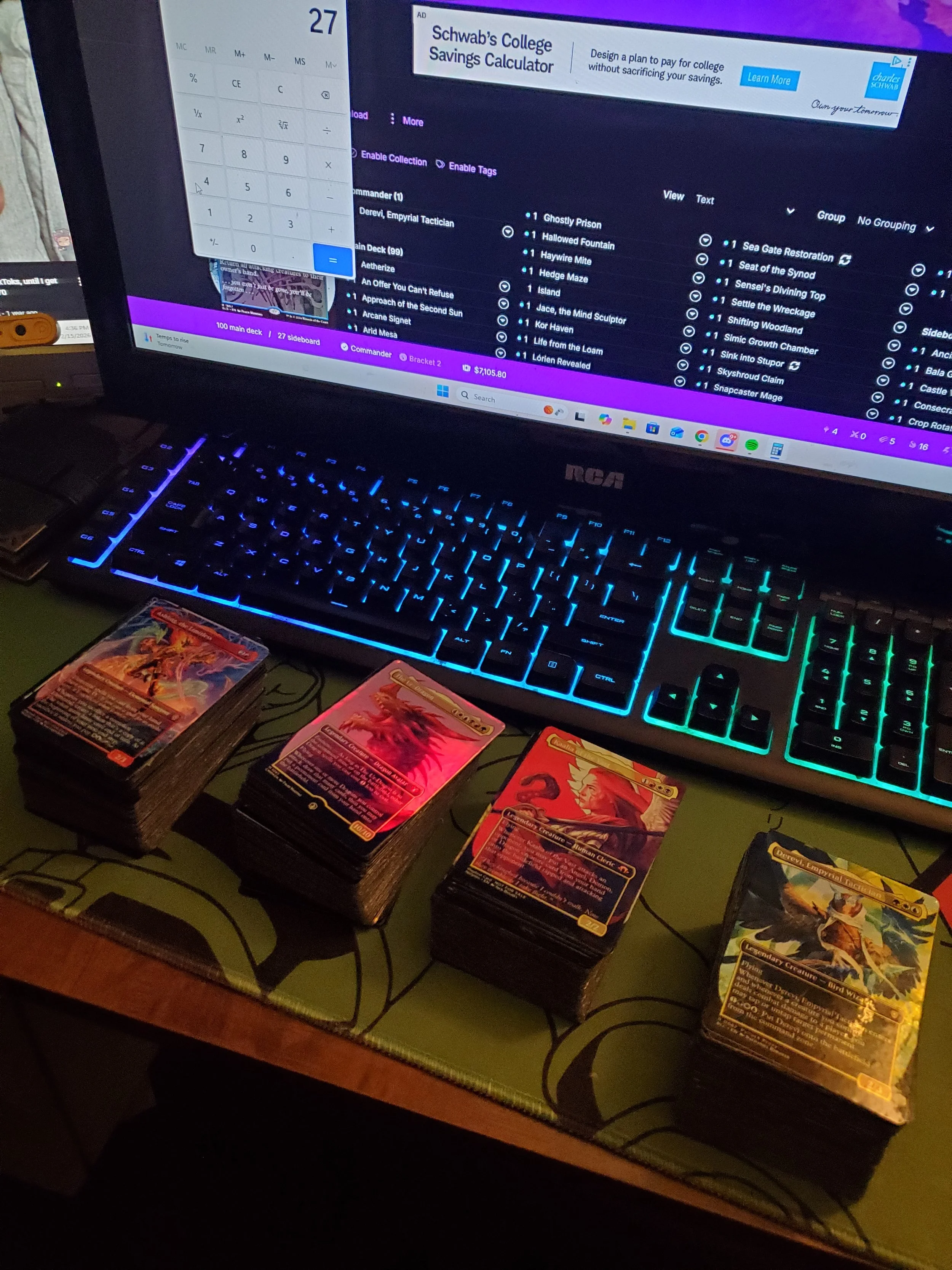 A computer monitor displaying a digital card game deck management screen, with a backlit keyboard, and four stacks of trading cards lined up on a desk in front of it.