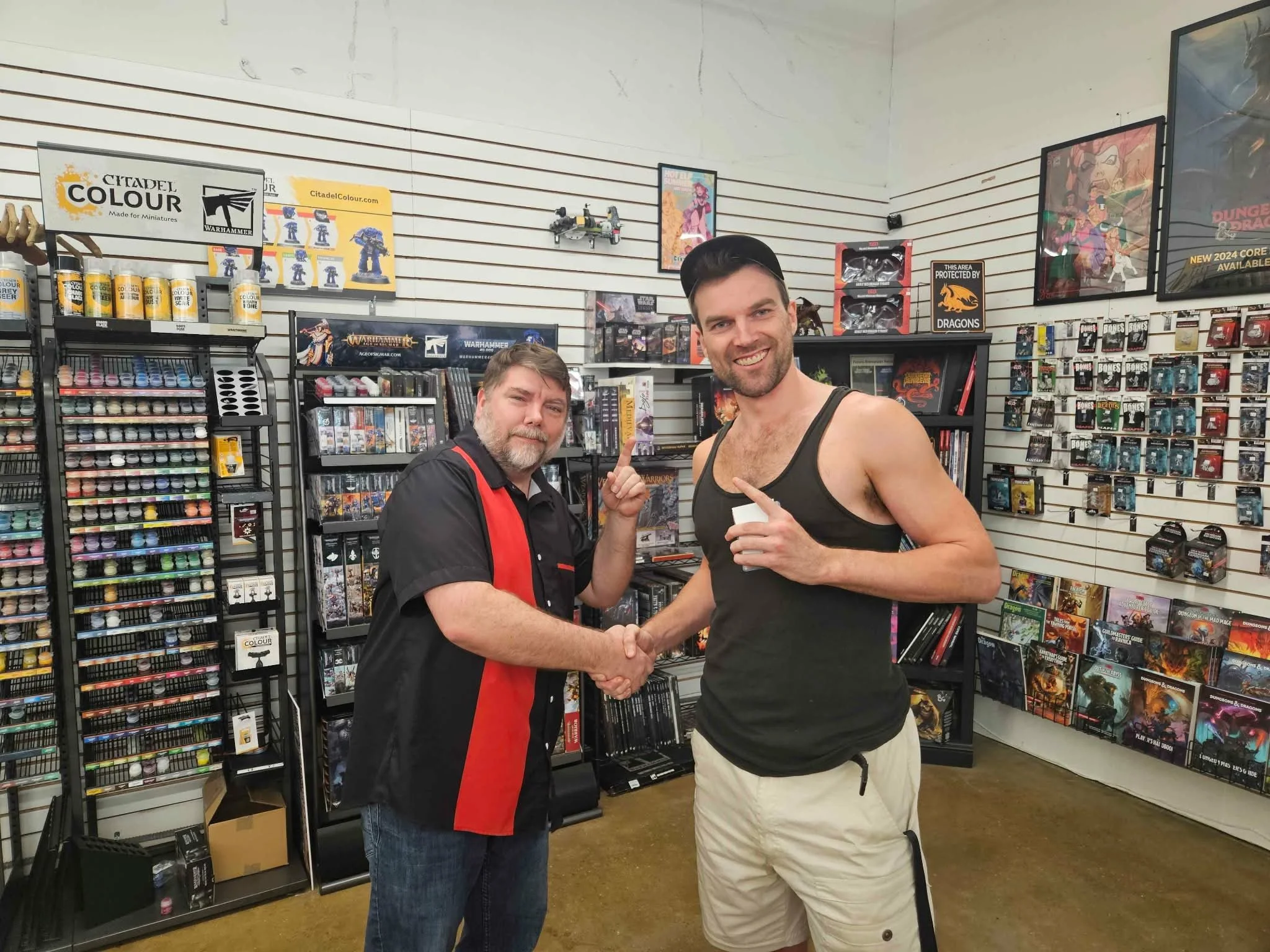 Two men shake hands inside a gaming store with shelves of miniature paints, games, and posters, one wearing a black and red shirt, the other in a black tank top and beige shorts.