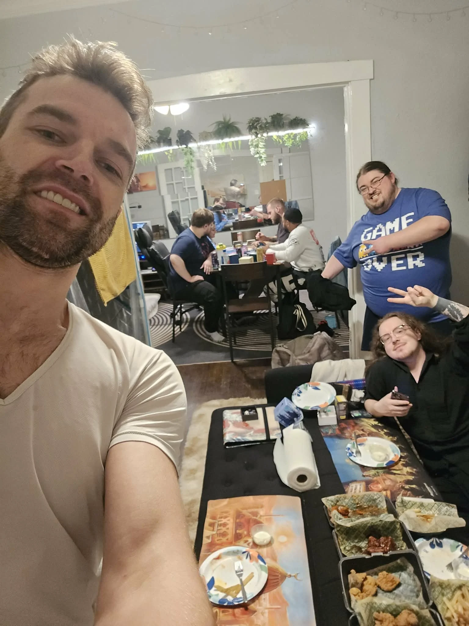 Selfie of a group of friends at a dinner or gaming gathering, with food and drinks on the table, some people sitting and some standing, in a room with a large mirror and plants.