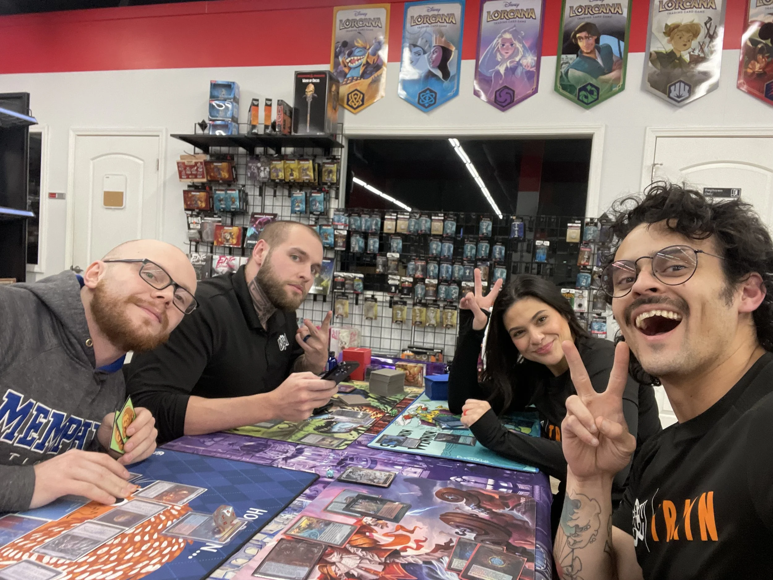 Four friends playing a card game at a store specializing in trading card games, with shelves of cards and colorful banners hanging on the wall.