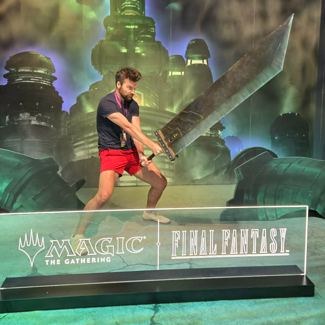 A man with a beard and curly hair wearing a black T-shirt and red shorts, holding a large sword with both hands in a pose. He is standing in front of a backdrop depicting a futuristic cityscape with tall buildings. A transparent sign in front of him reads 'Magic: The Gathering' and 'Final Fantasy'.