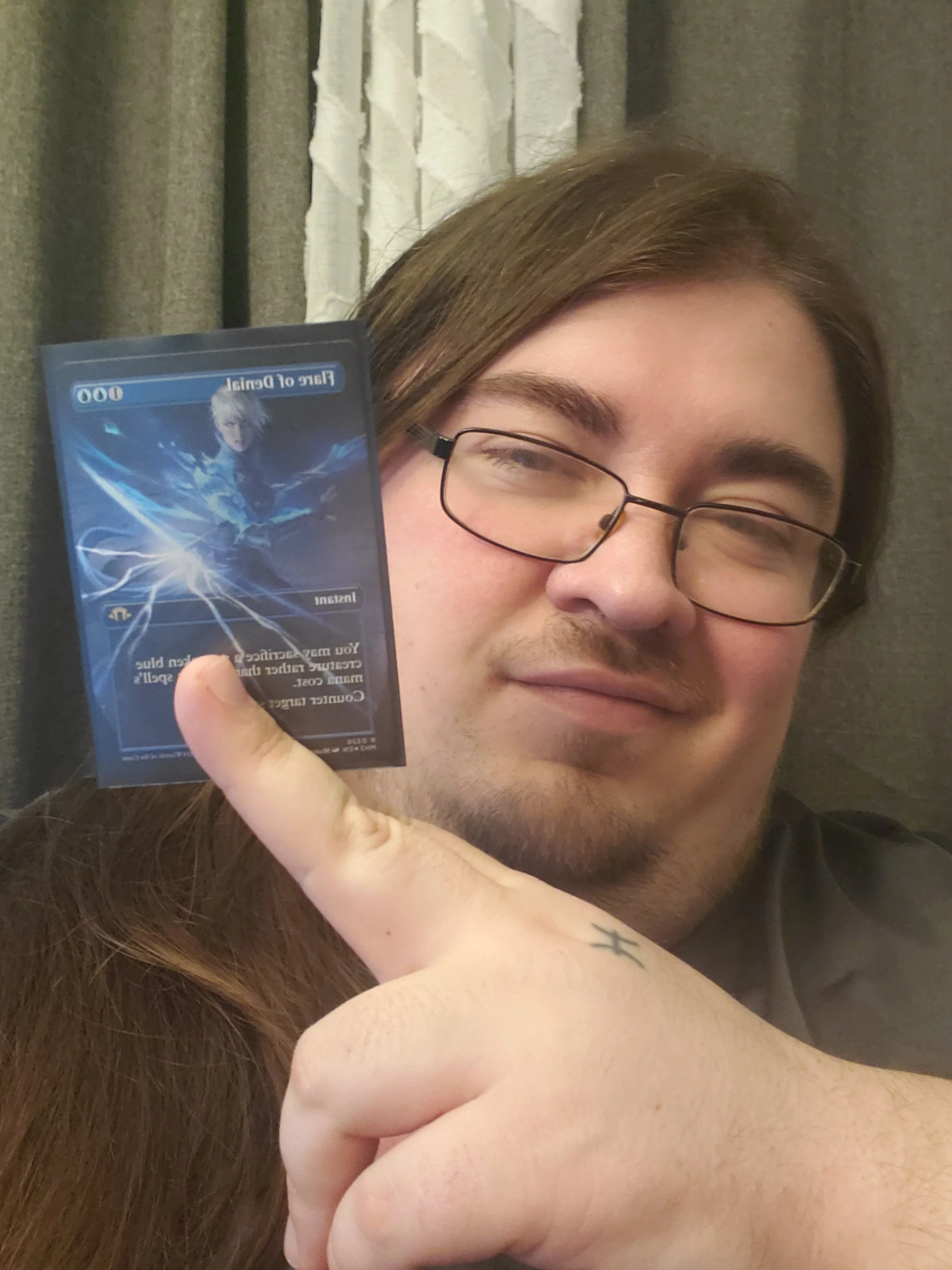Person with glasses holding a trading card featuring a character from a fantasy or sci-fi game or show, with curtains in the background.