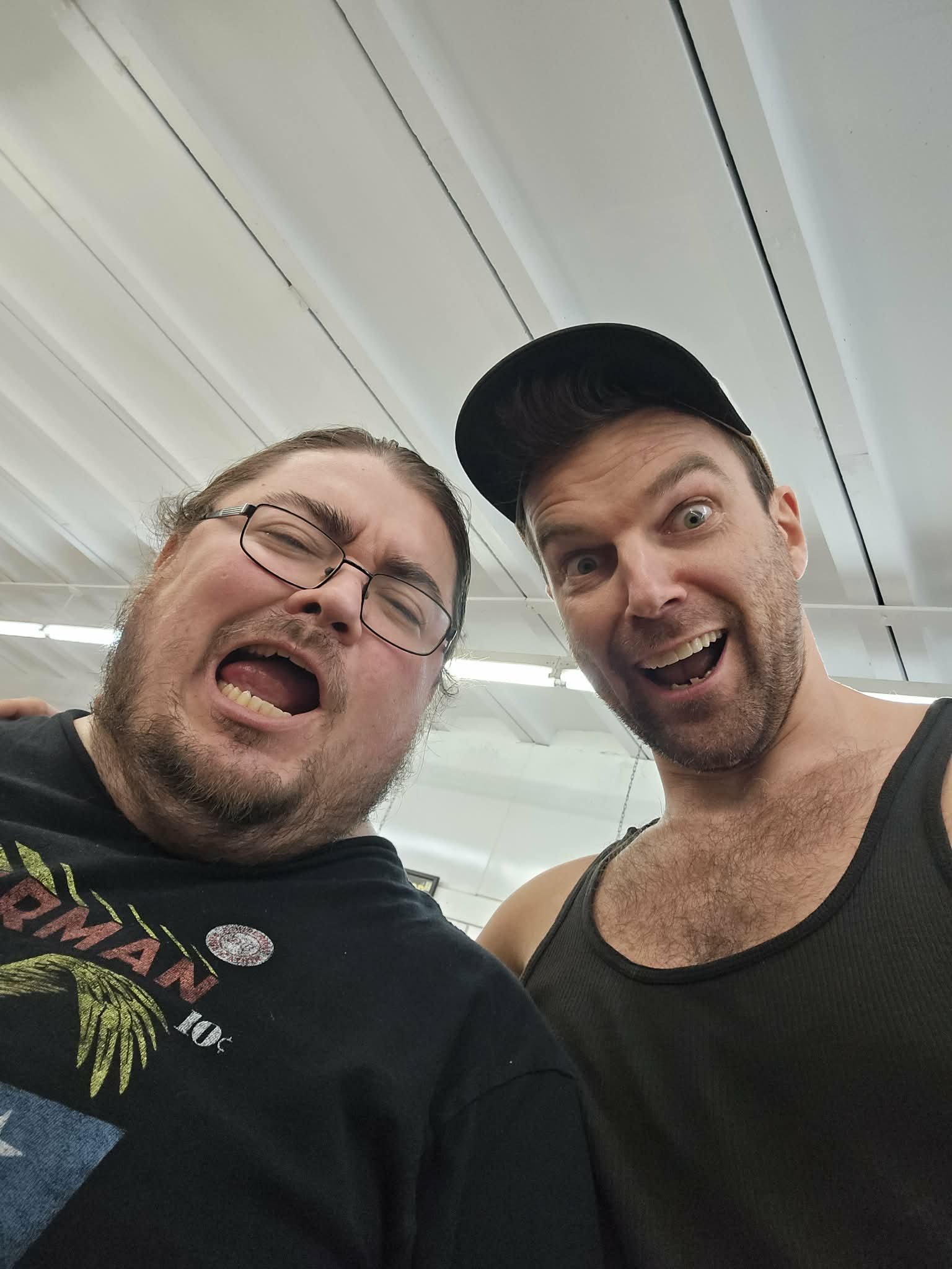 Two men taking a selfie together indoors, smiling and making expressive faces. One man is wearing glasses and a black graphic T-shirt, the other is wearing a black tank top and a black cap.