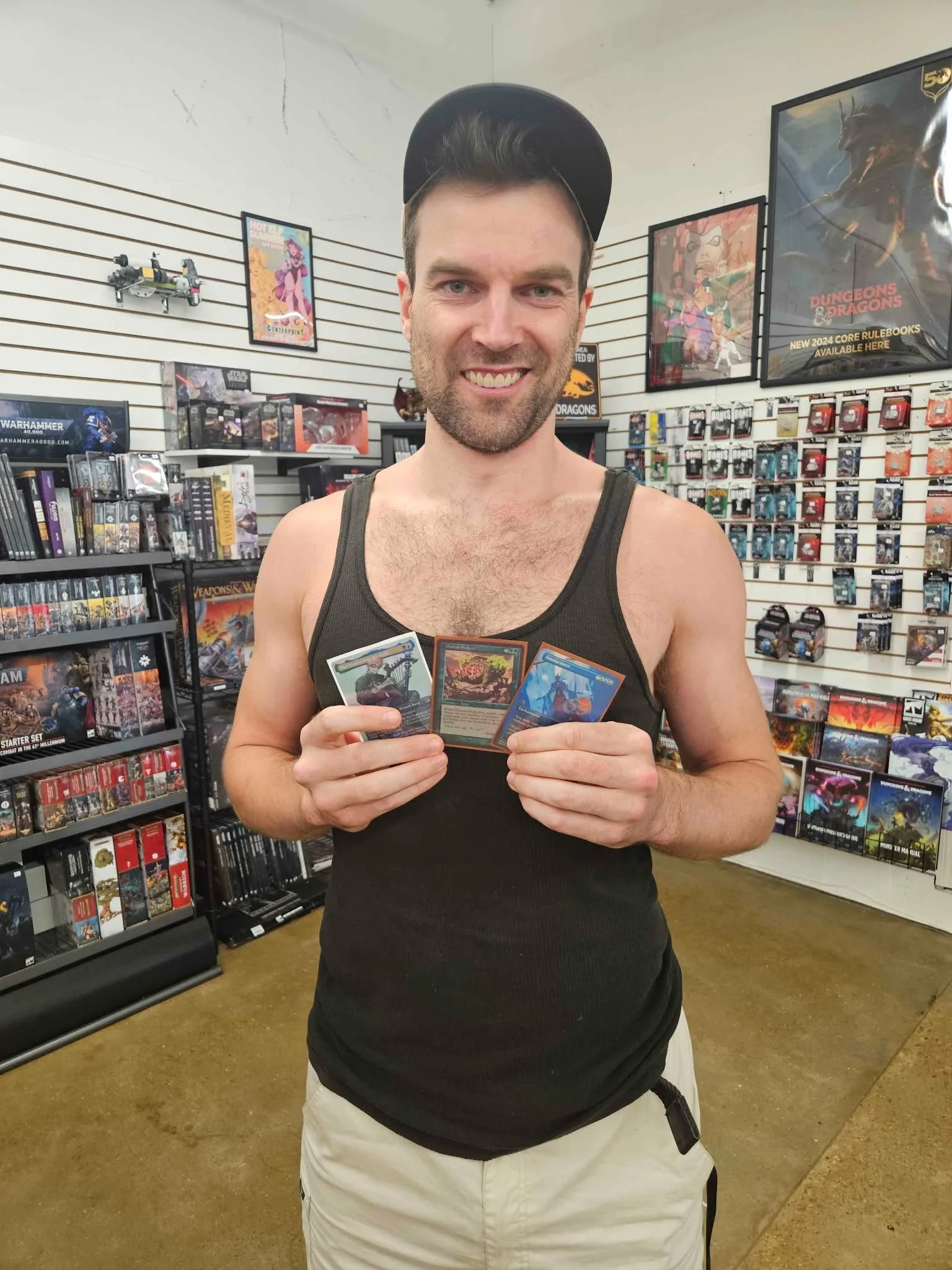 A young man in a black tank top and baseball cap is smiling and holding four colorful trading cards in a game shop filled with board games, trading cards, and posters.