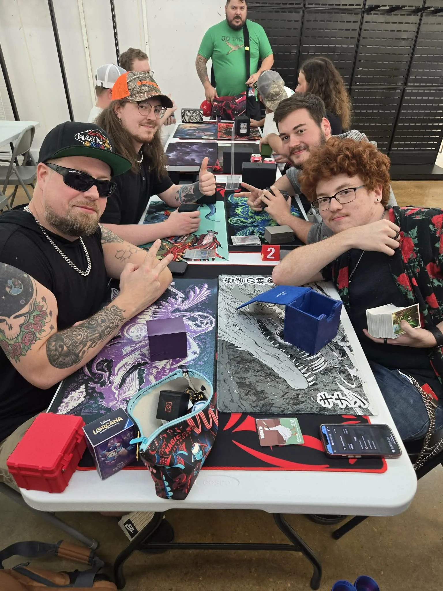 A group of people sitting around a table playing card and board games, with some giving thumbs up. There are gaming accessories and cards on the table, and a man standing in the background.