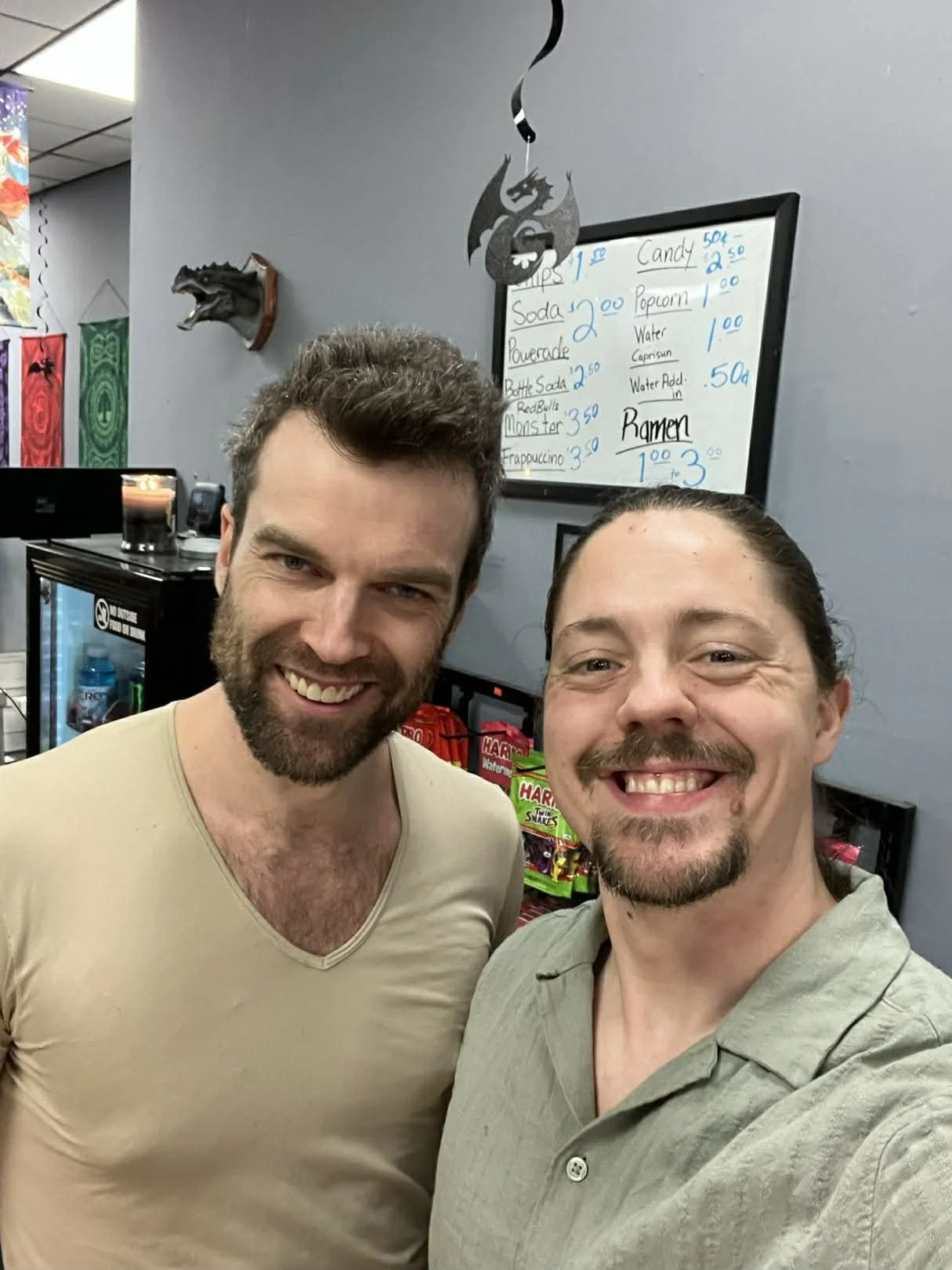 Two smiling men taking a selfie in a store or food stand, with a menu board listing drink and snack prices in the background.