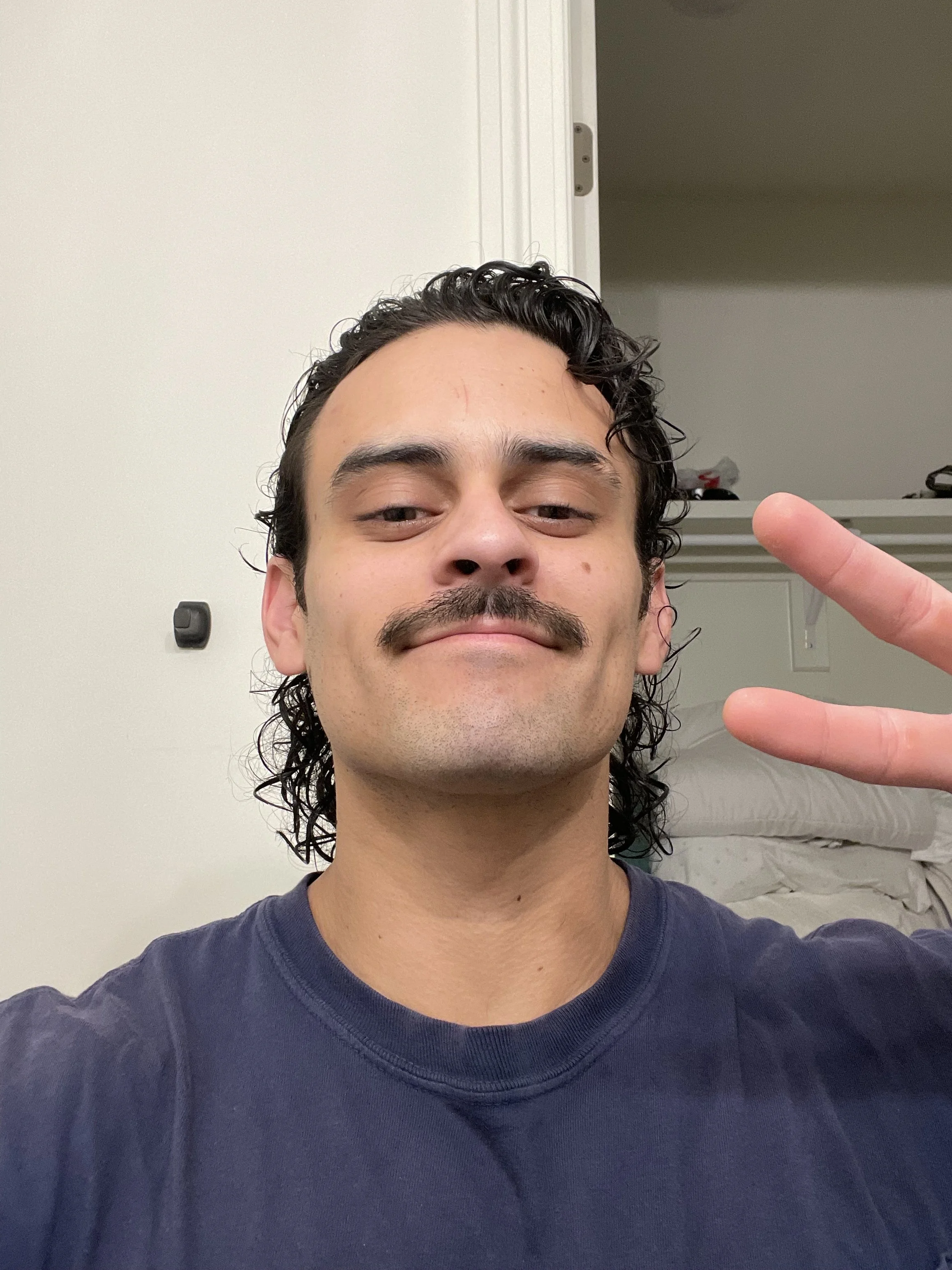 A young man with dark curly hair and a mustache taking a selfie, making a peace sign with his right hand, standing in a room with a white door and closet with shelves in the background.