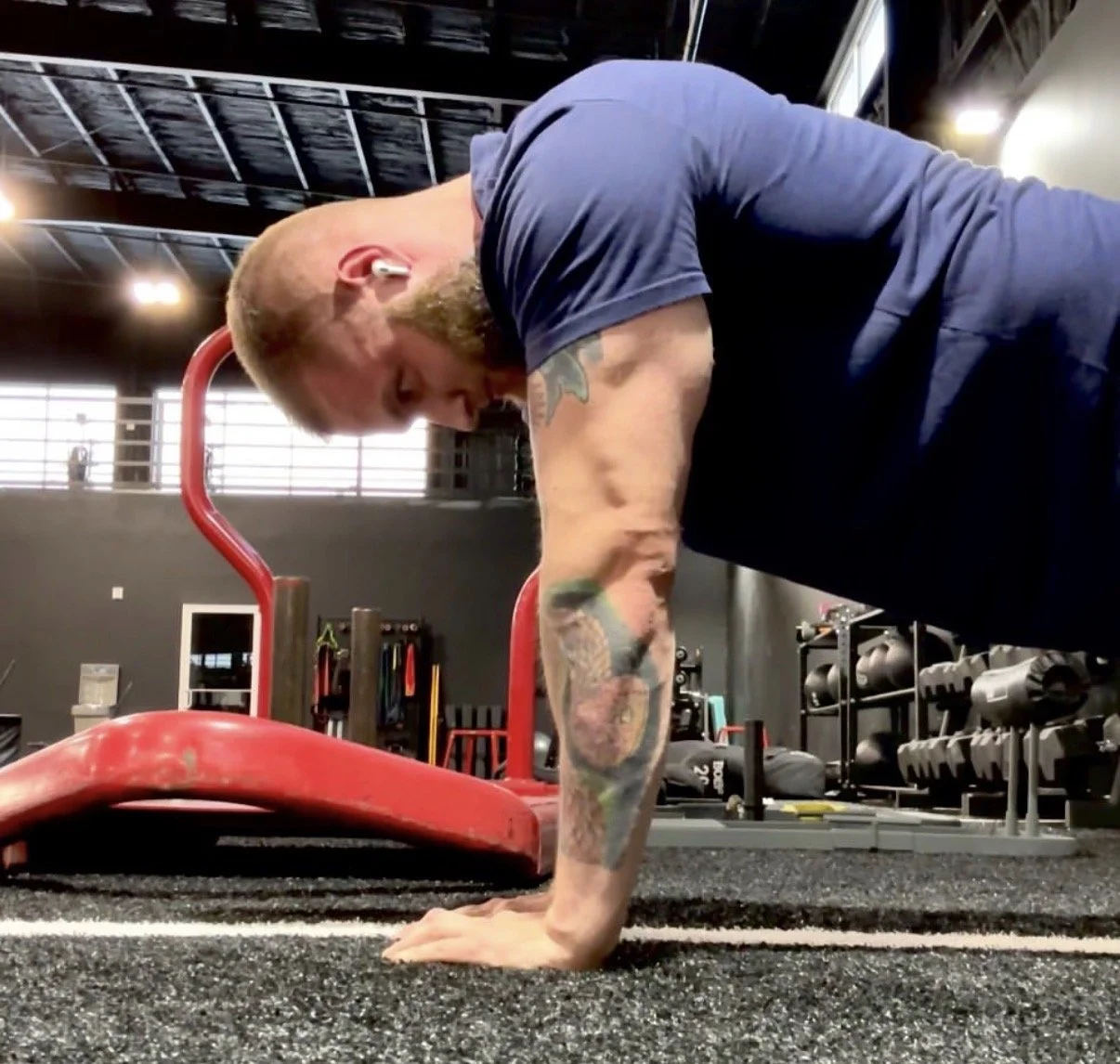 A man in a gym doing a push-up on the floor, wearing wireless earbuds, a navy blue shirt, and showing tattoos on his left arm.