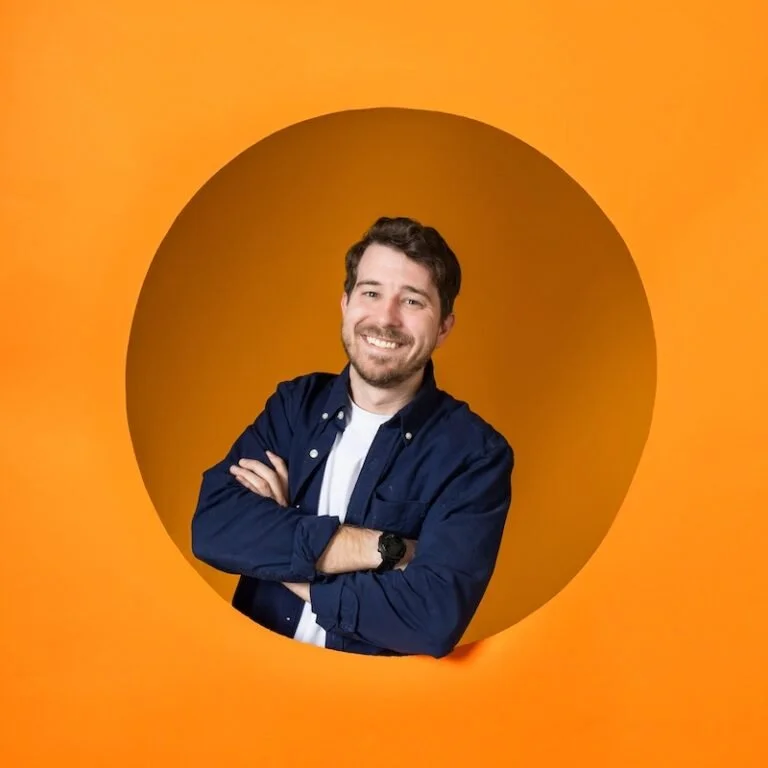 Man wearing blue button up and smiling with his arms crossed while leaning through a large circular opening in a vibrant orange backdrop.