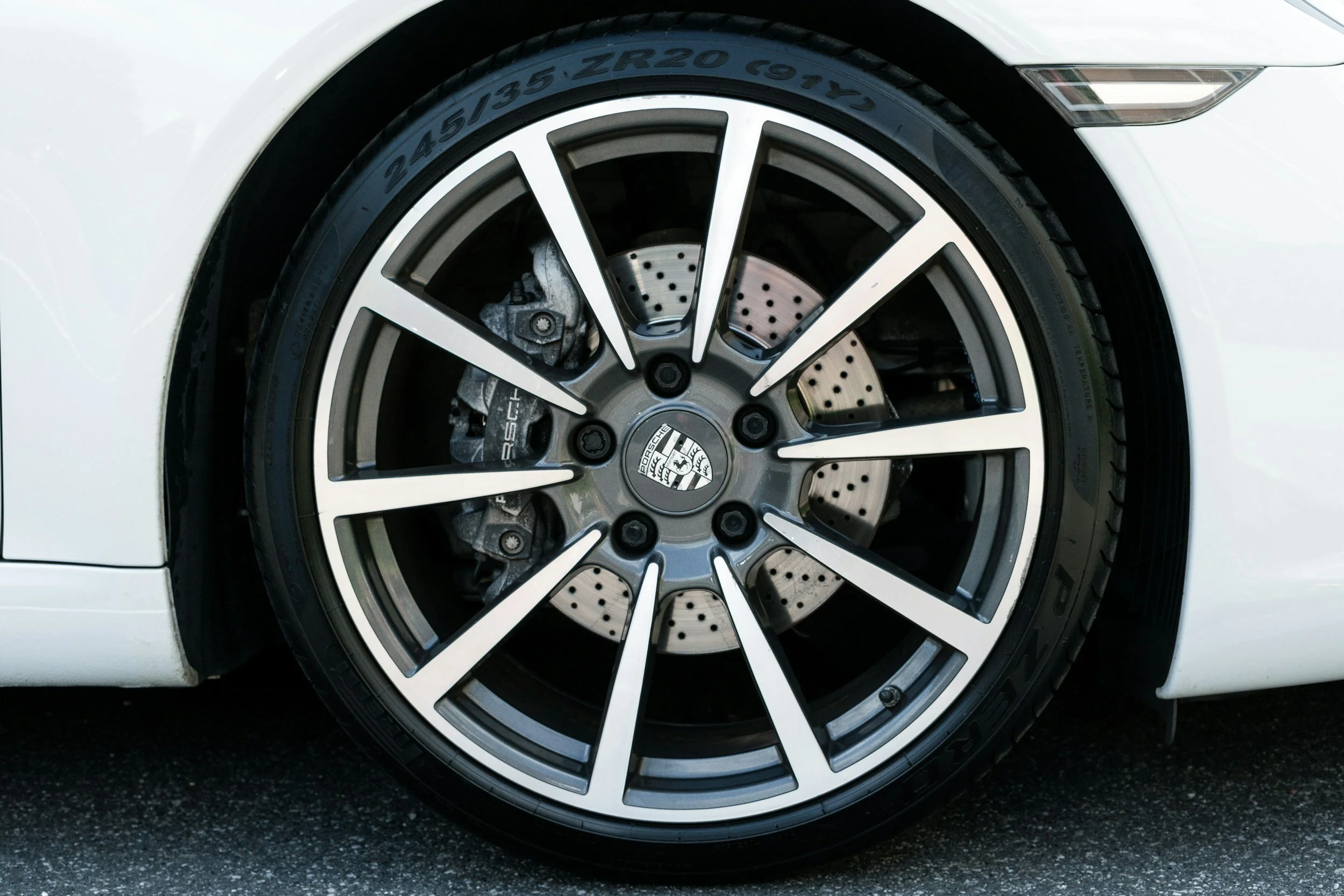 Close-up of a luxury car wheel showing a black and silver alloy rim with a Porsche logo at the center, exposed brake rotors, and a black tire with visible size markings.