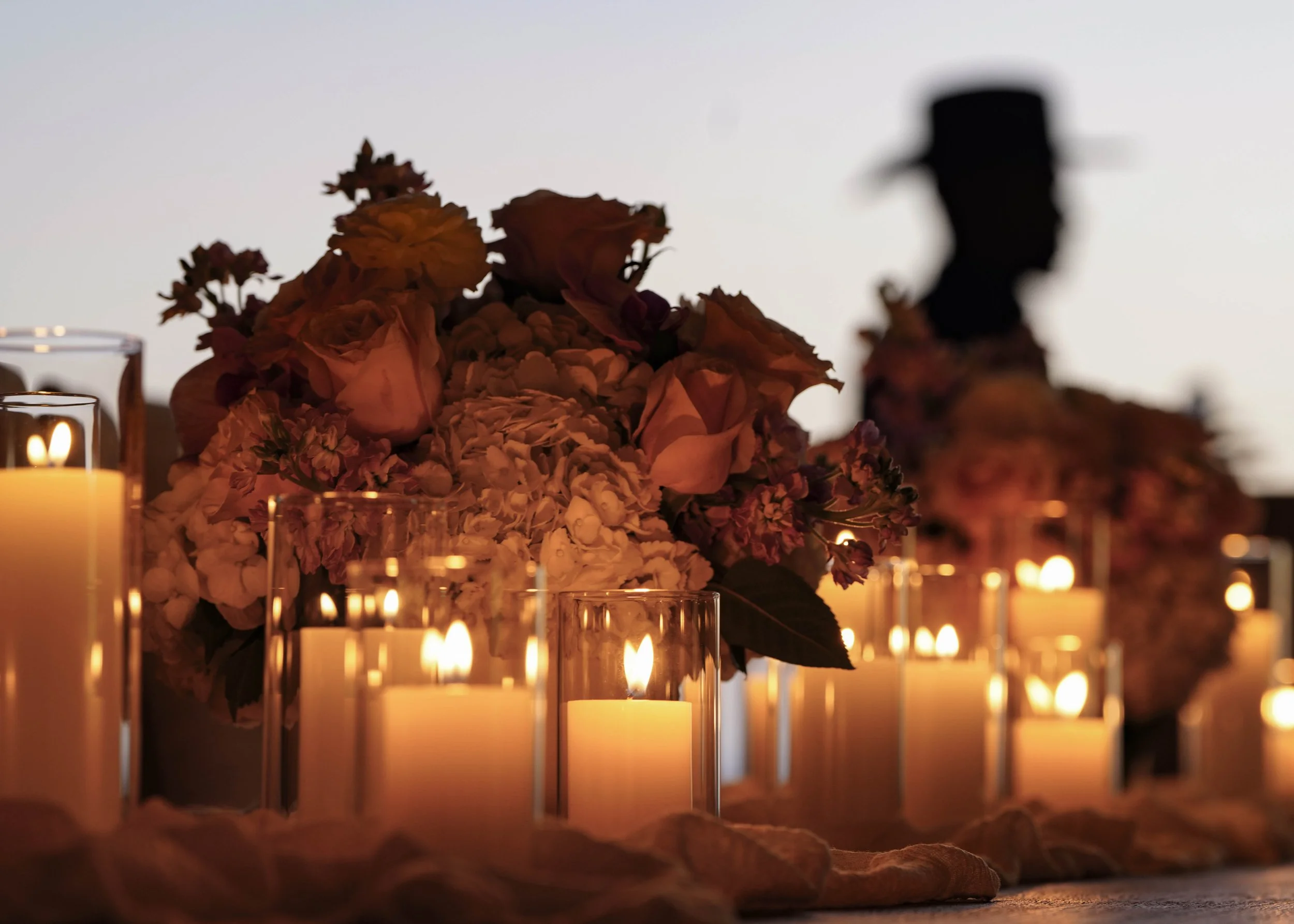 Photo by Black Light Imaging - Close up of floral arrangement and lit candles on a table. Dark outline of a person walking behind the table.