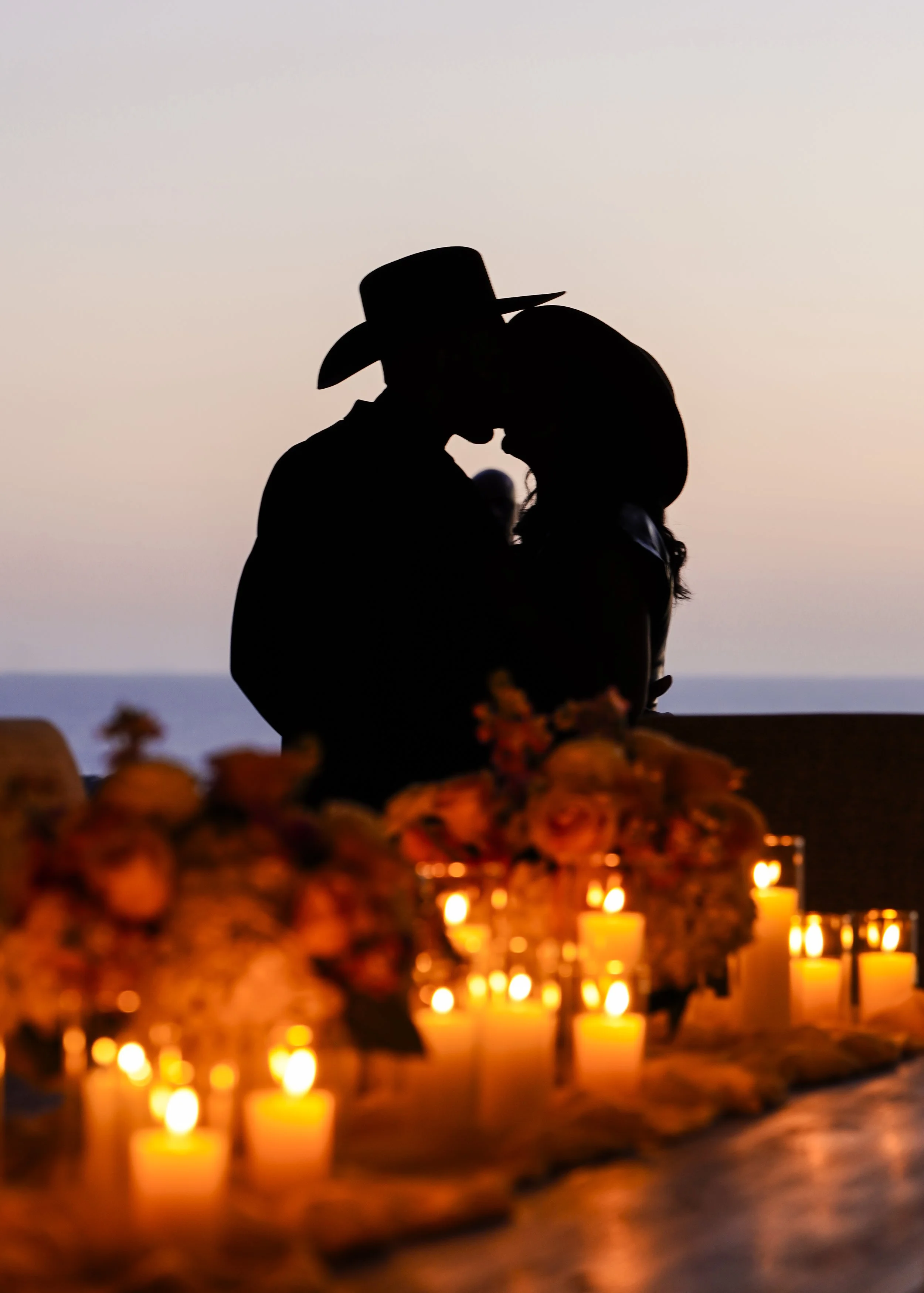 Photo by Black Light Imaging - Couple sharing a kiss as the sunsets over the ocean. Table with flower arrangements and lit candles in front of them.