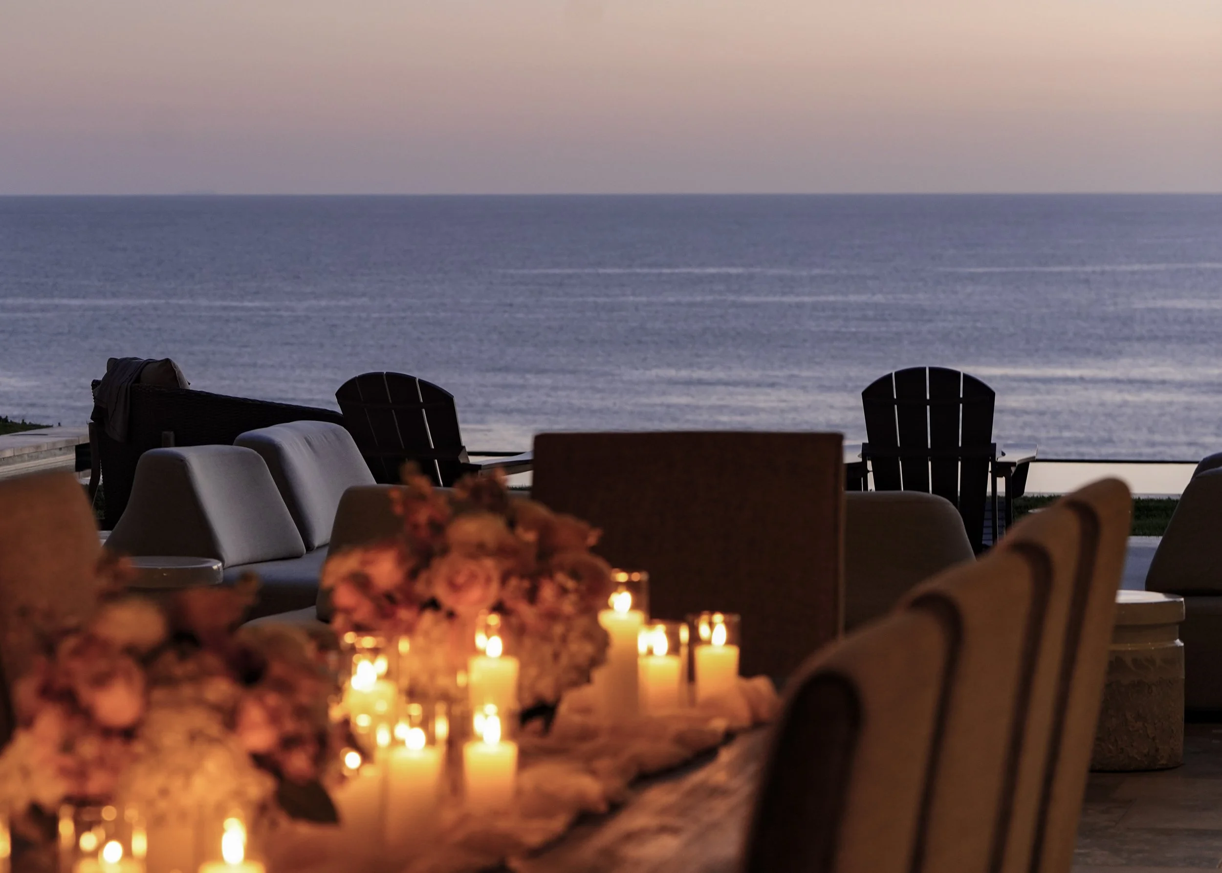 Photo by Black Light Imaging - Table with floral arrangements and lit candles alongside outdoor chairs on a patio overlooking the ocean during sunset.