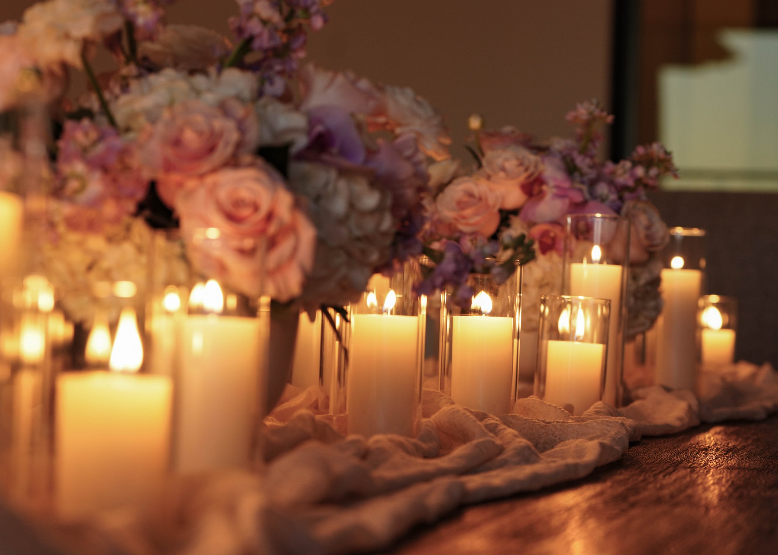 A romantic table setting with multiple lit candles in glass holders and large pink, purple, and white flower arrangements.