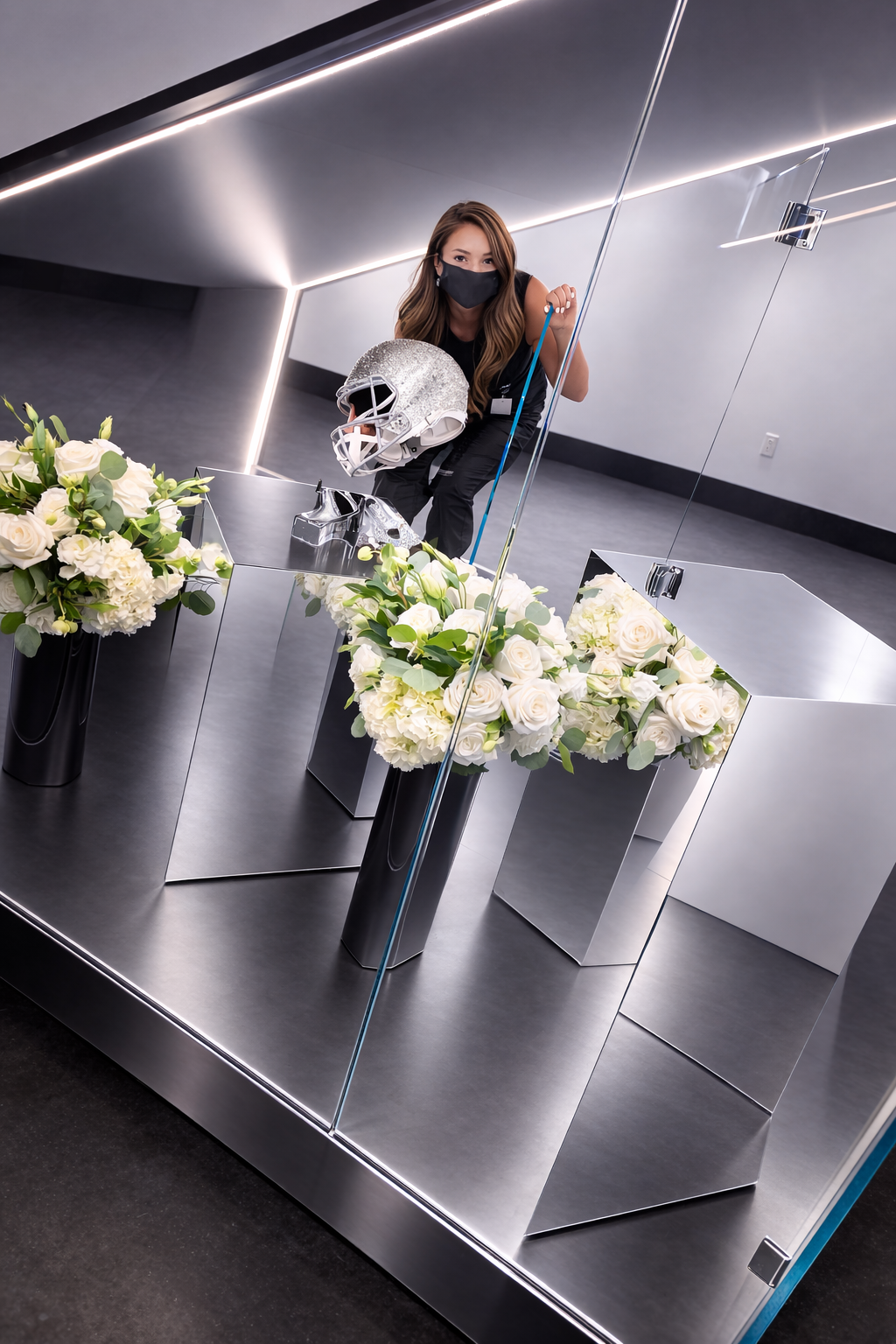 A woman wearing a black face mask holds a sparkling silver football helmet while looking into a mirror at a modern, metallic display of white roses and greenery in a sleek, contemporary setting.