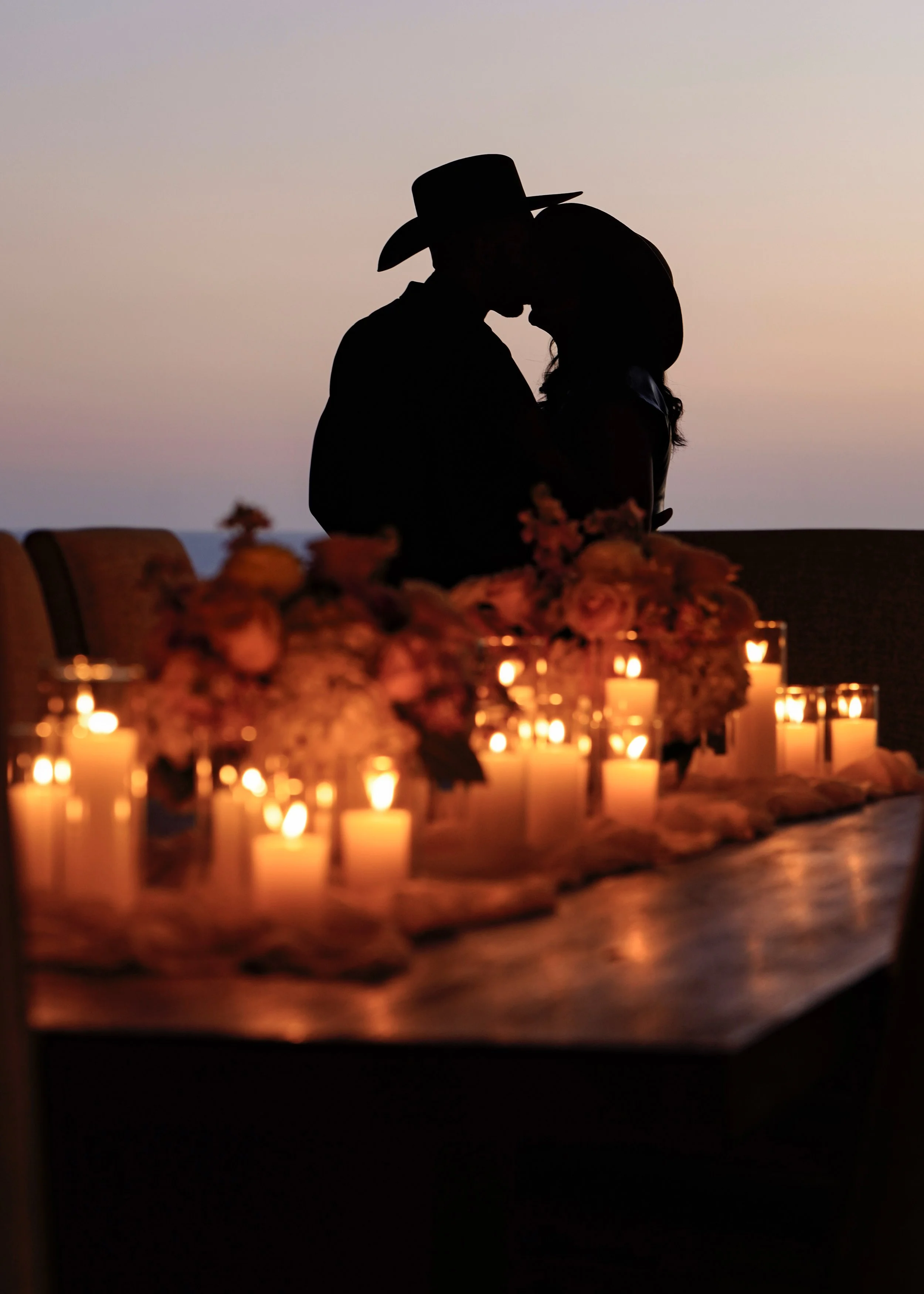 Silhouette of a couple wearing cowboy hats, embracing at a table decorated with candles and flowers during sunset.