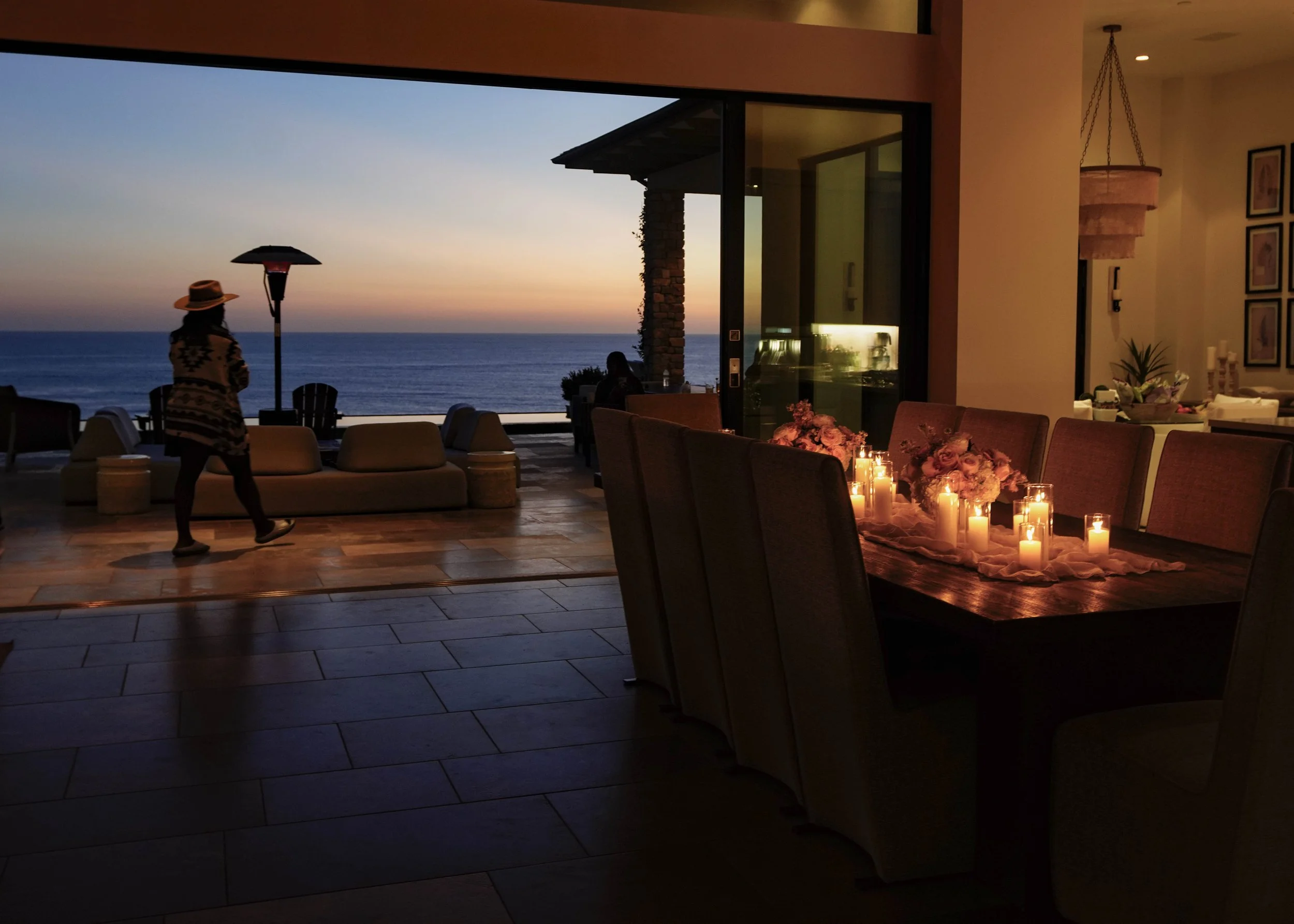 Photo by Black Light Imaging - Woman walking across patio overlooking the ocean during sunset. Table with floral arrangements and lit candles on right side of her.